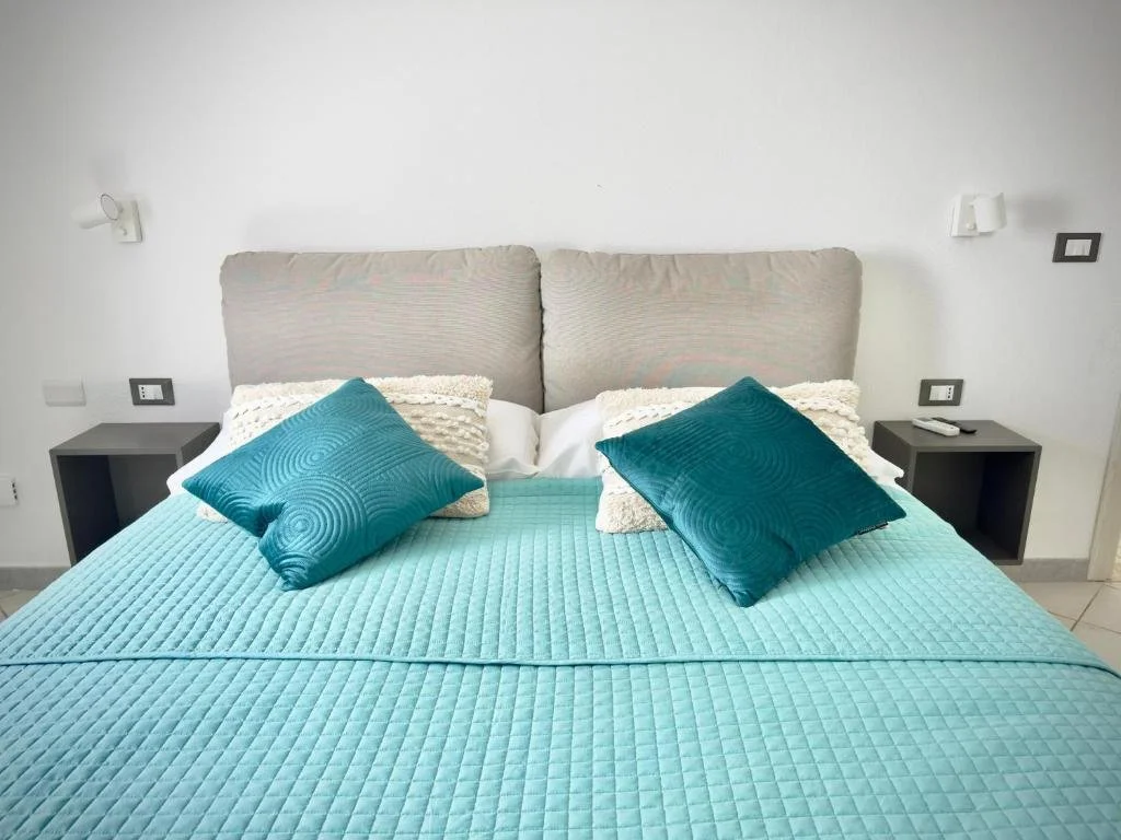 A neatly made bed with a light blue quilt, two dark blue decorative pillows, and three white textured pillows, flanked by white wall-mounted reading lights and small dark-colored nightstands with a remote control on one.