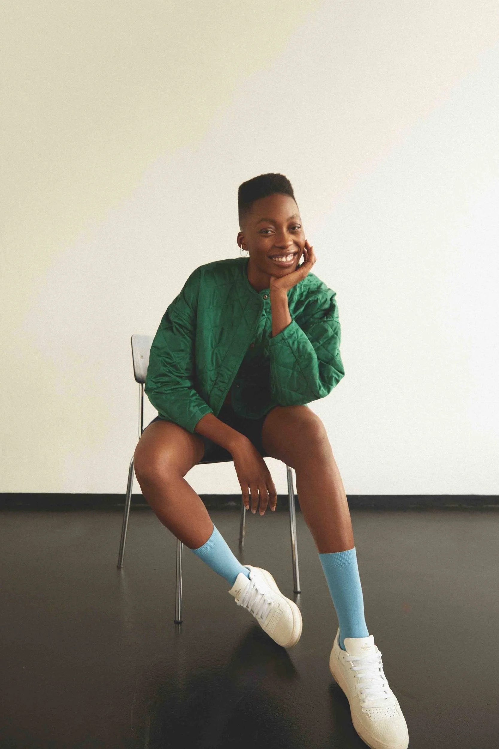 Green knit jacket, seated on a chair with blue socks and white sneakers