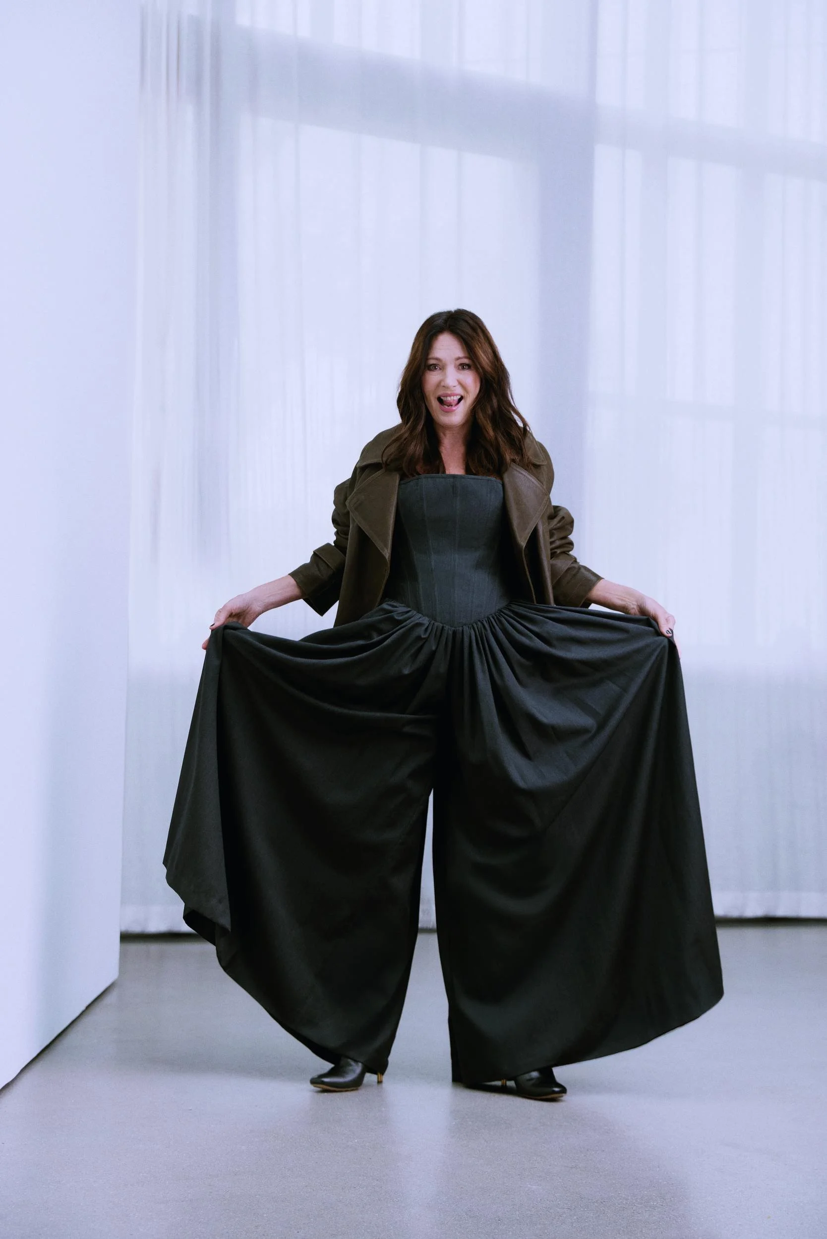Iris Berben holding out wide black palazzo trousers in front of a white curtain