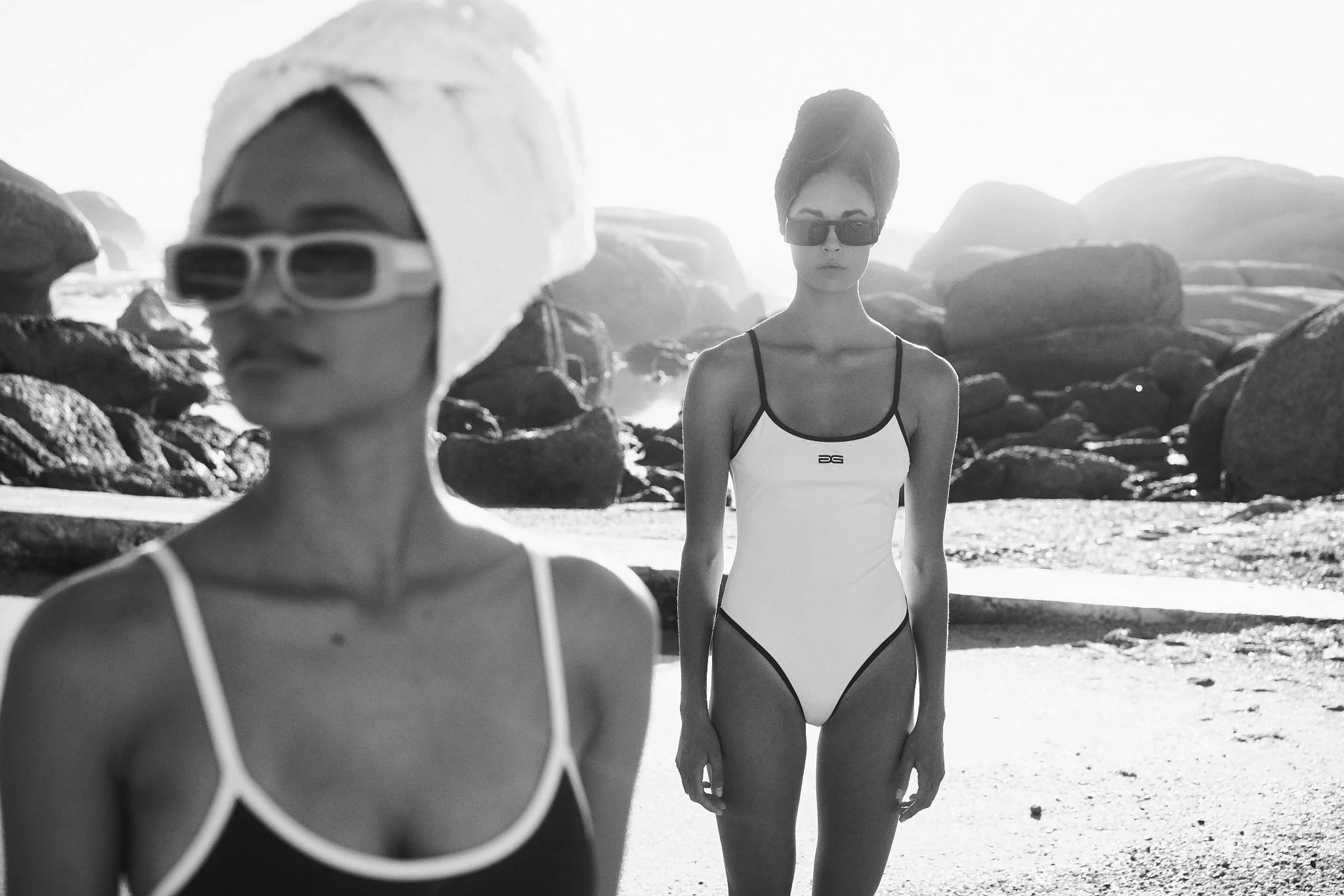 Black and white shot of two models in white and black one-piece swimsuits wearing towel turbans and sunglasses, boulders in the background