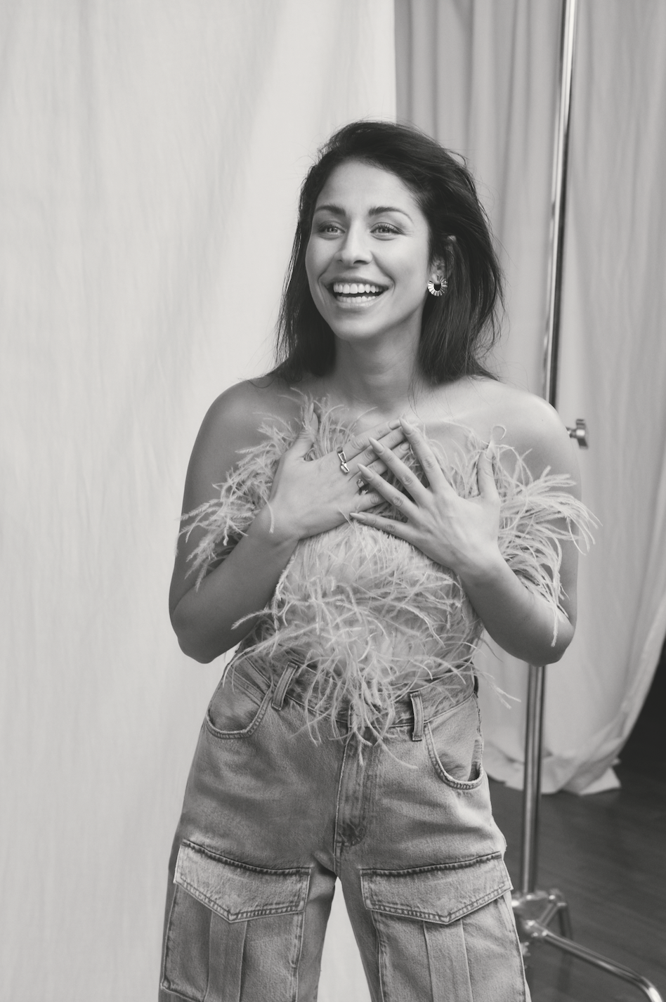 Black and white portrait laughing in a feathered top and denim cargo trousers, hands crossed on chest