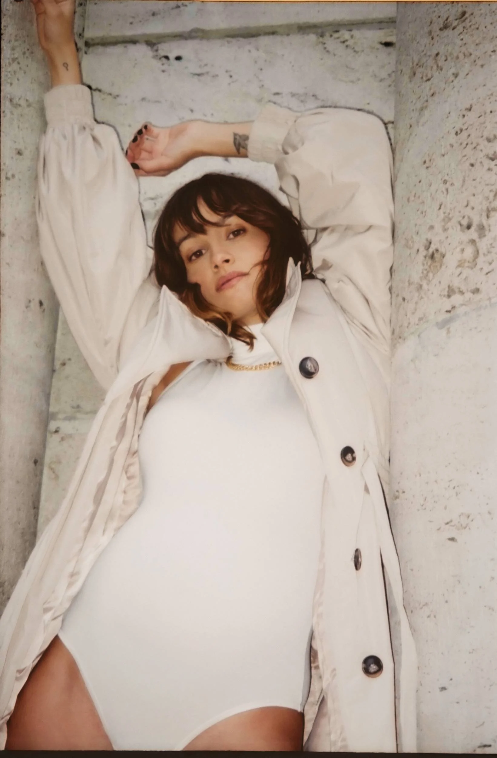 Mogli in a white bodysuit and cream coat lying on stone steps