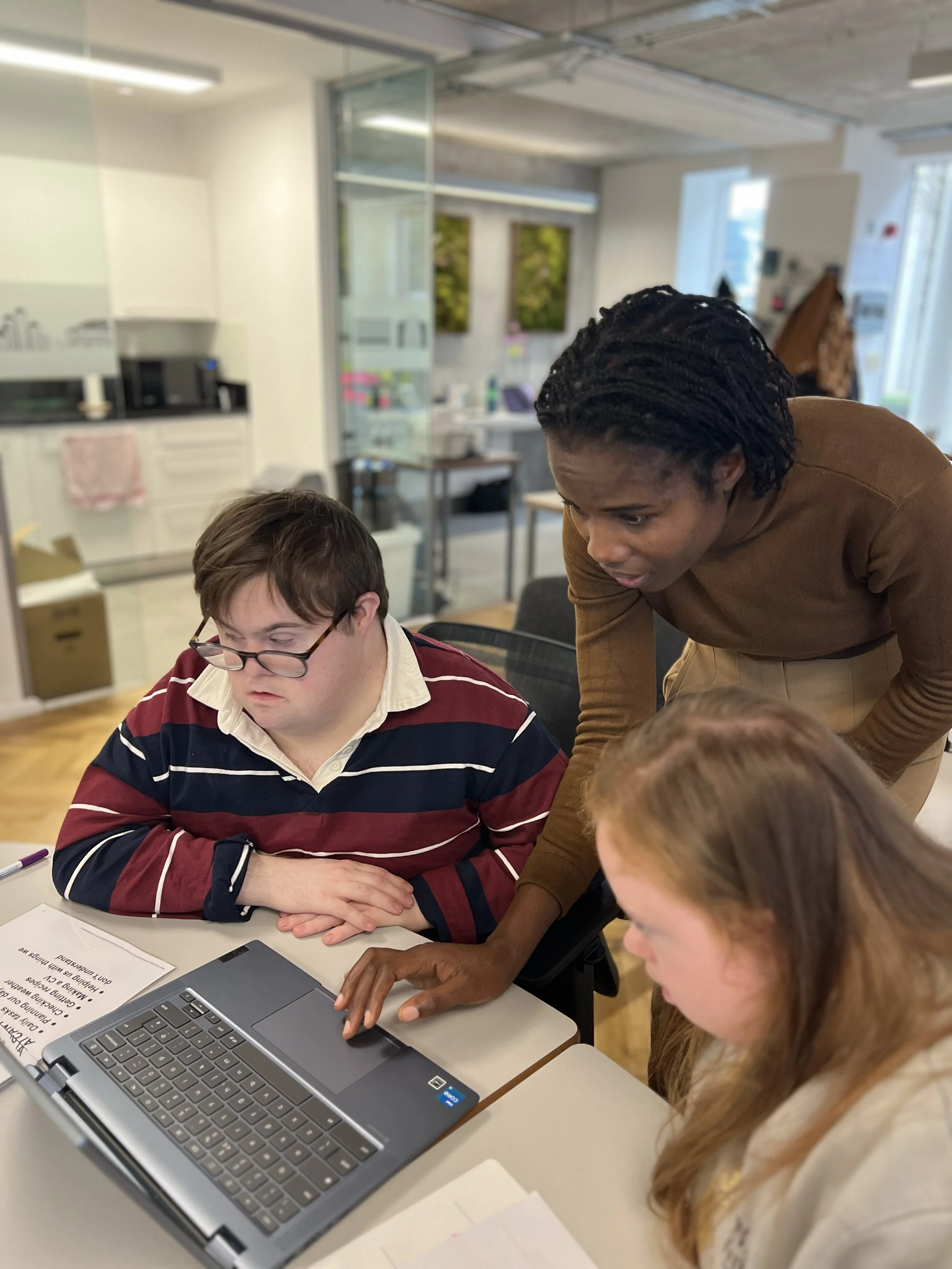 Three young adults gathered around a laptop, smiling and engaging in a discussion in an office setting.