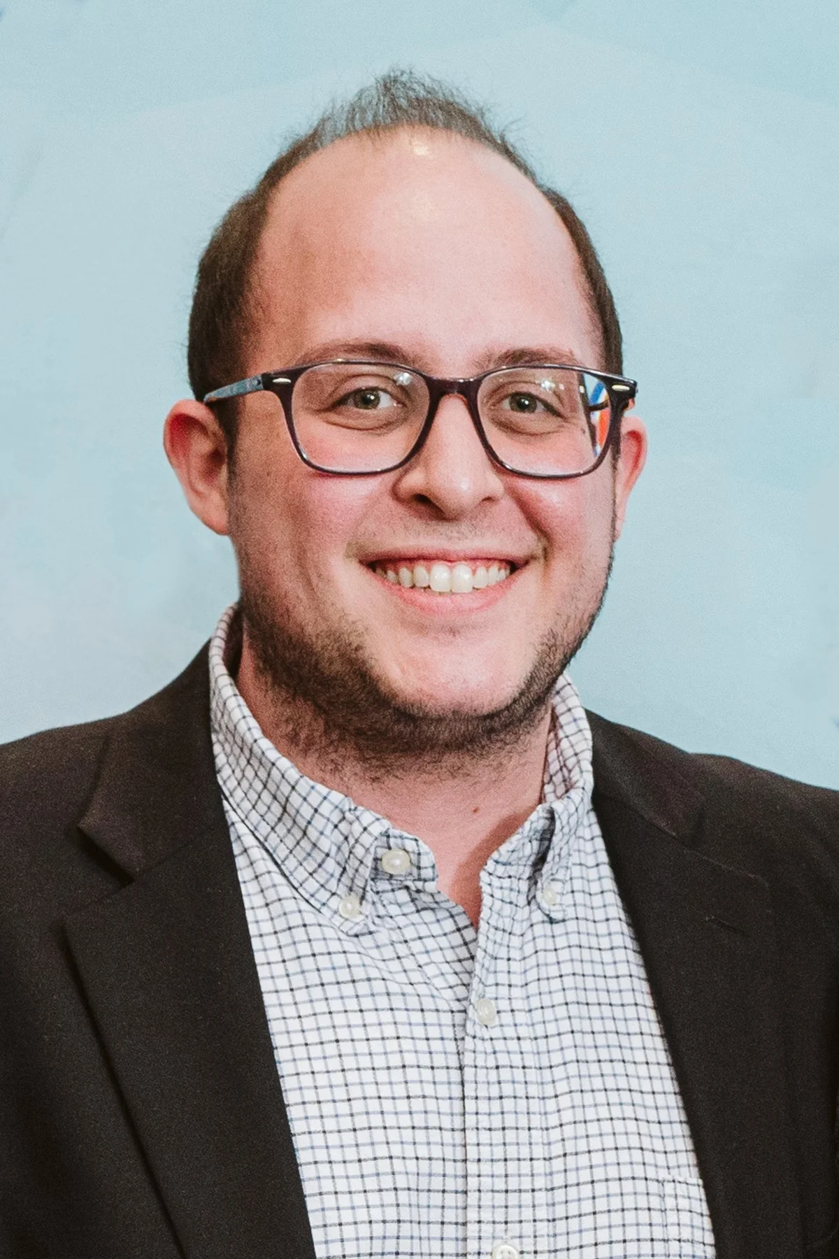 Smiling person wearing glasses, a plaid shirt, and a dark blazer.