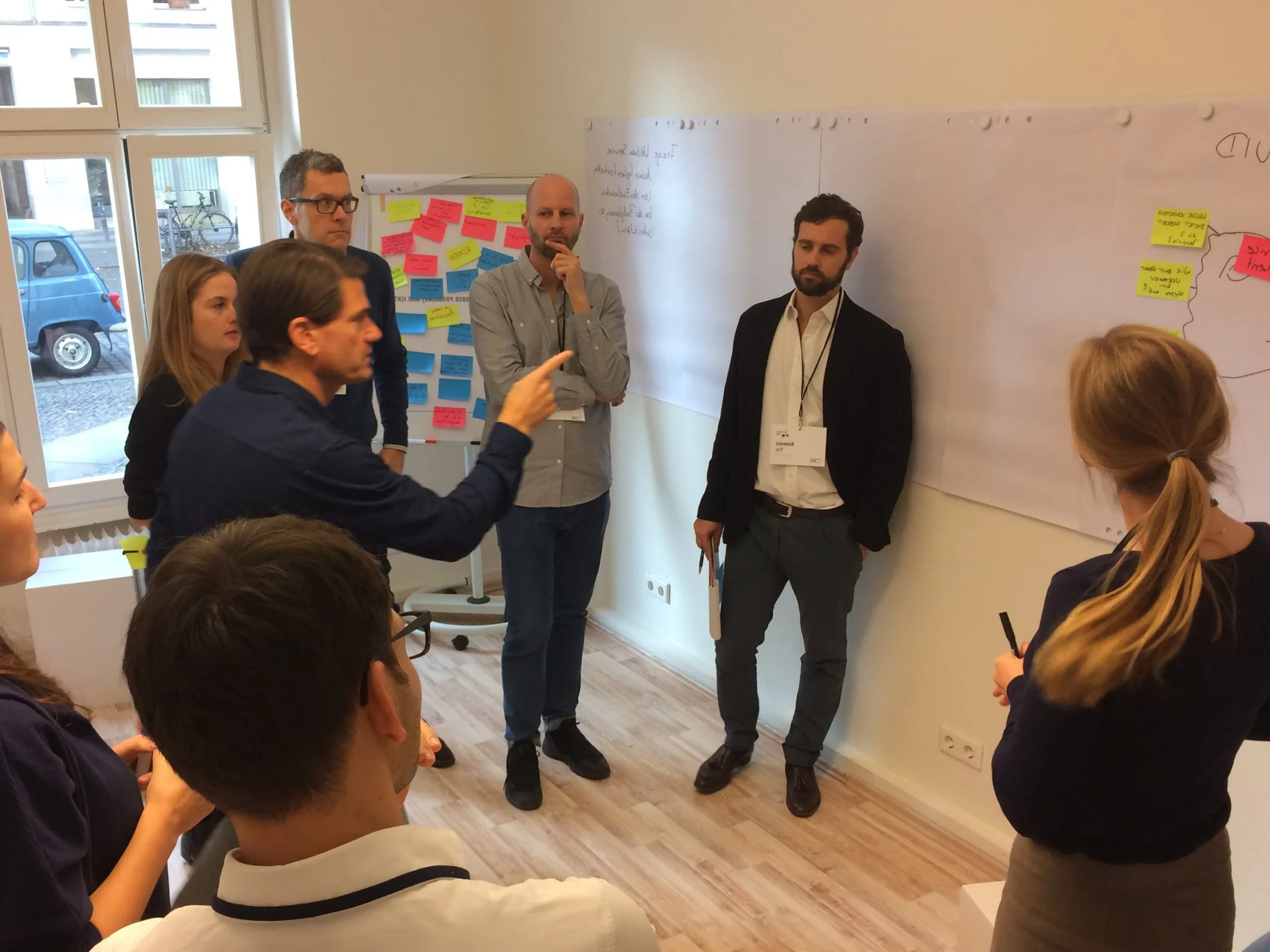 A workshop managed by Alessio Tixi, with people engaged in speaking, gesturing, while others are listening attentively. It's a group activity supported by a whiteboard with sticky notes and other office related preps.