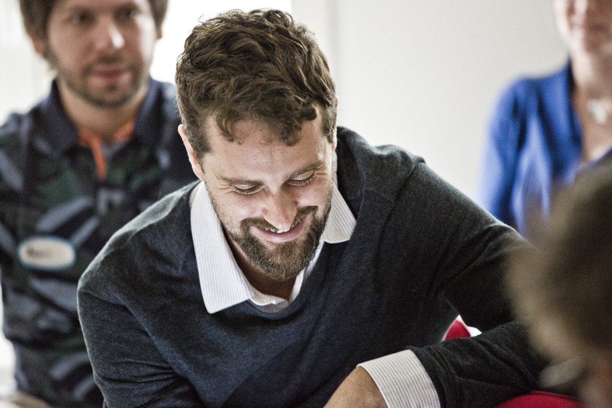 Alessio Tixi smiling while looking down, engaged in conversation, with blurred people in the background.