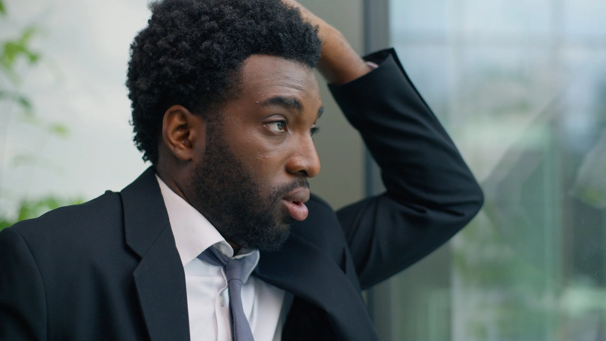 A man in a black suit and tie looking out a window with a worried or contemplative expression, resting his head on his hand.