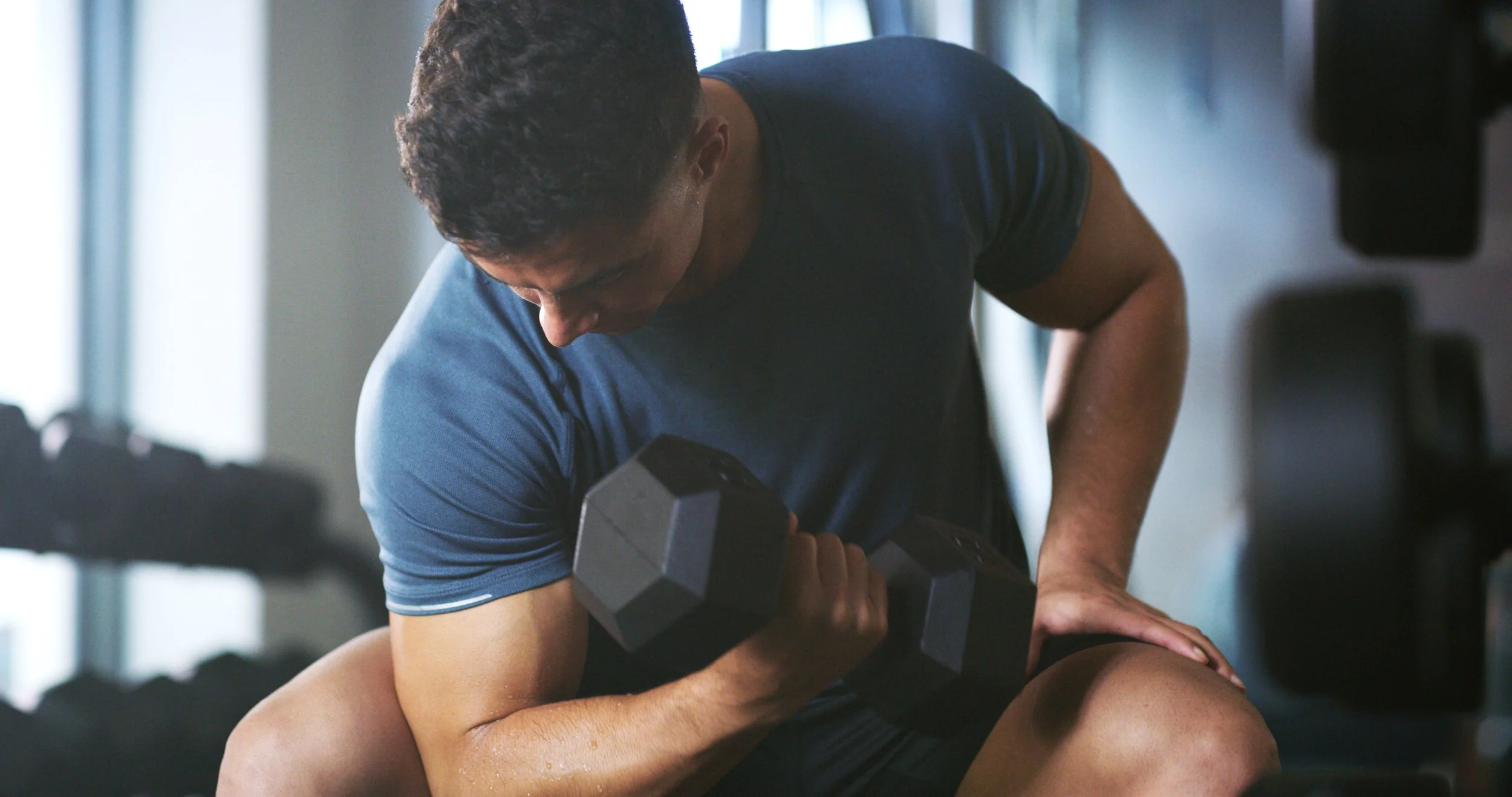 A man in a blue shirt lifting a dumbbell while seated and embracing a woman who is lying on his lap in a gym.
