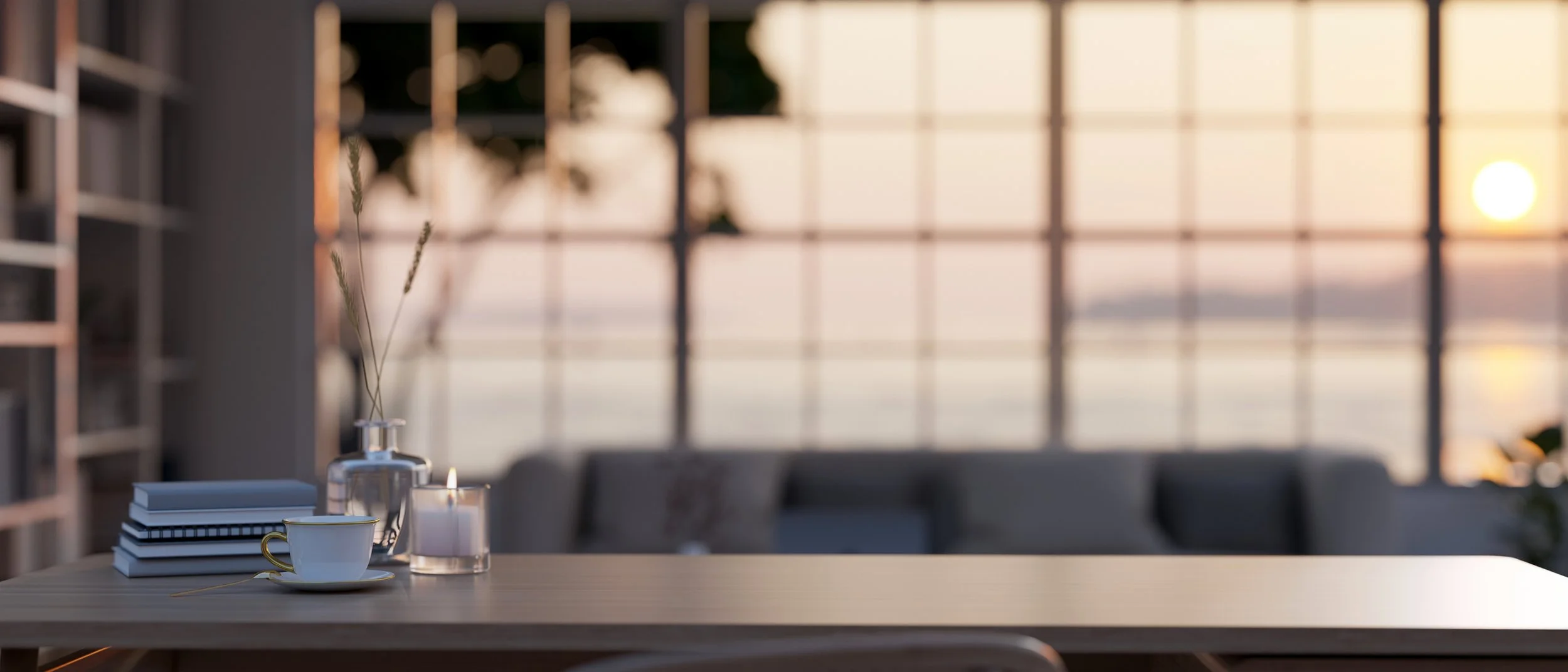 A wooden table with a stack of books, a white coffee cup with a gold rim, a glass bottle with tall grass, and a candle, with a large window and a view of the sunset in the background.