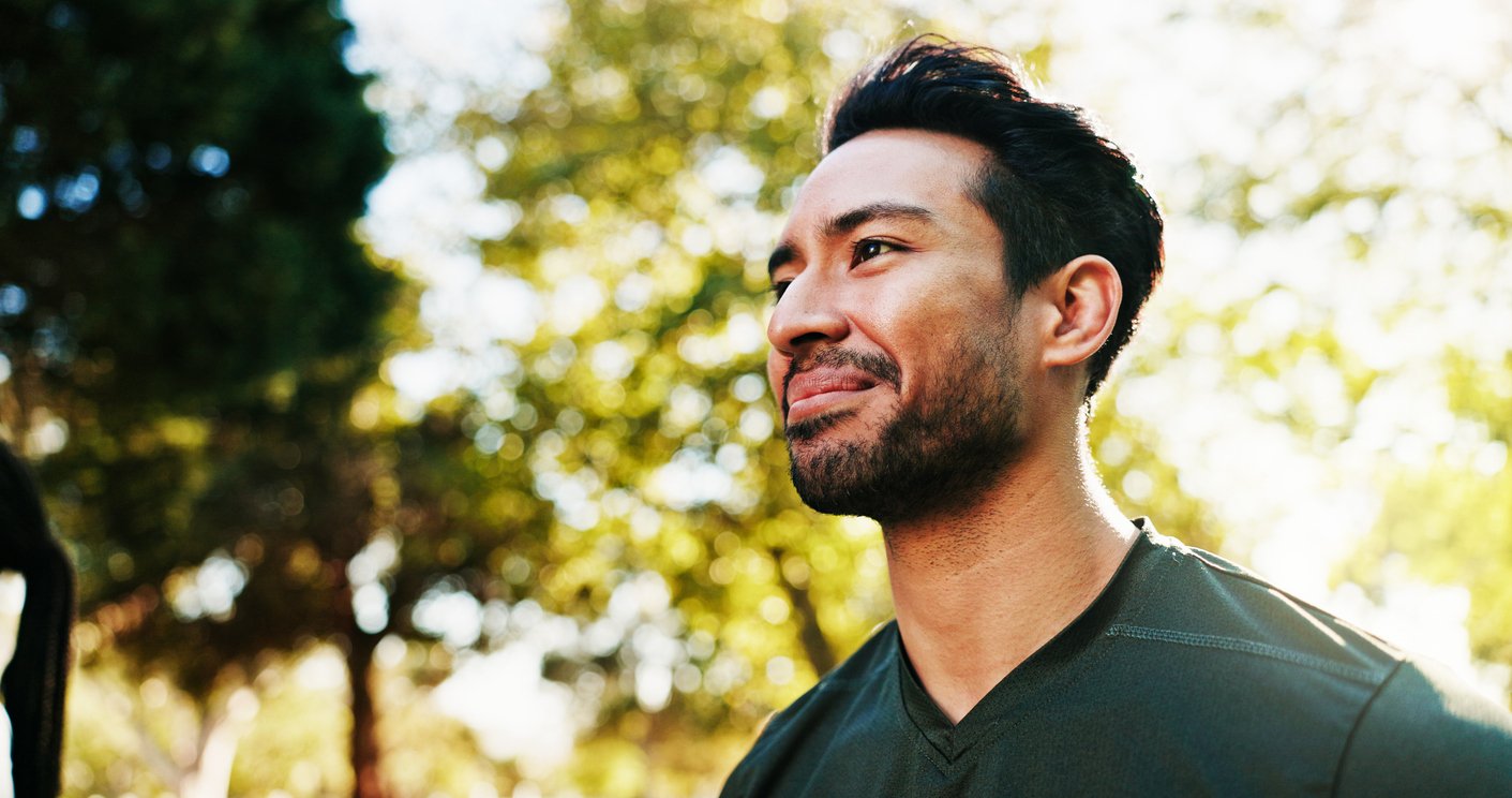 A man with dark hair and a beard outdoors with sunlight filtering through trees, looking to the side.