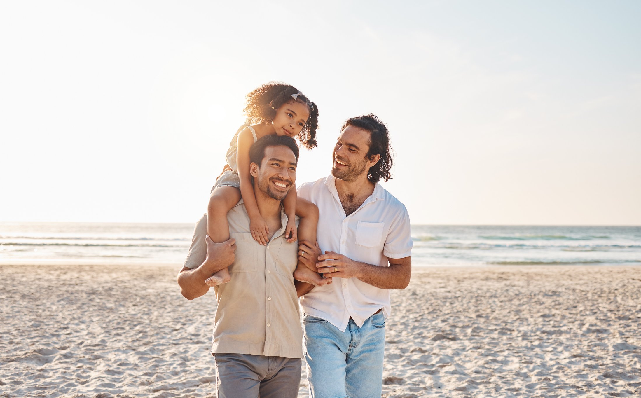 Two men walking on the beach with a young girl on one of their shoulders, all smiling and enjoying a sunny day by the ocean.