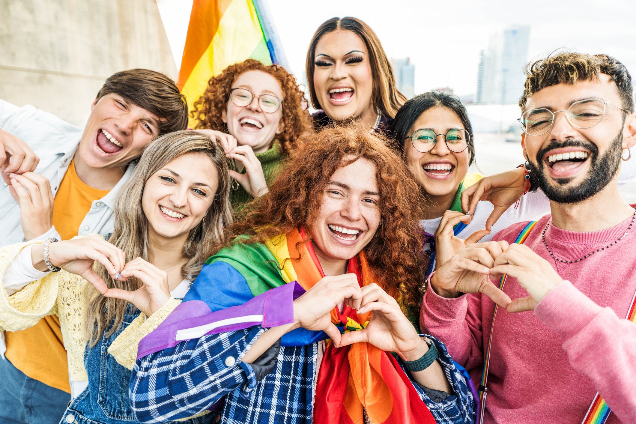 Group of diverse young people smiling, making heart gestures, and celebrating with rainbow flags, indicating LGBTQ+ pride.