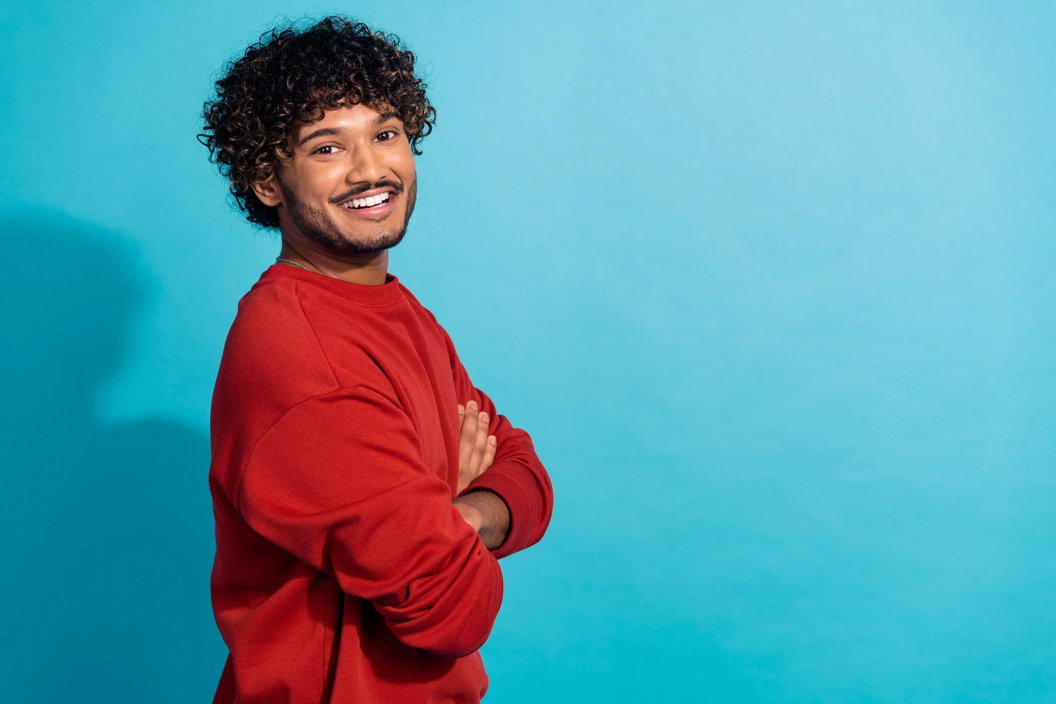 A young man with curly hair and a beard, wearing a red long-sleeve shirt, standing with arms crossed and smiling against a blue background.