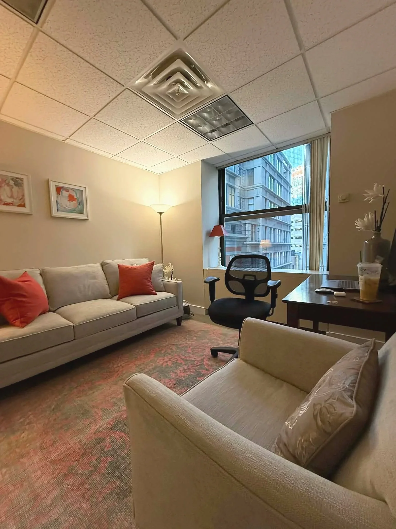 Interior of a hotel room furnished with a beige sofa with red pillows, an armchair, a black office chair, a wooden desk with a laptop and drinks, a floor lamp, and large window with city view.
