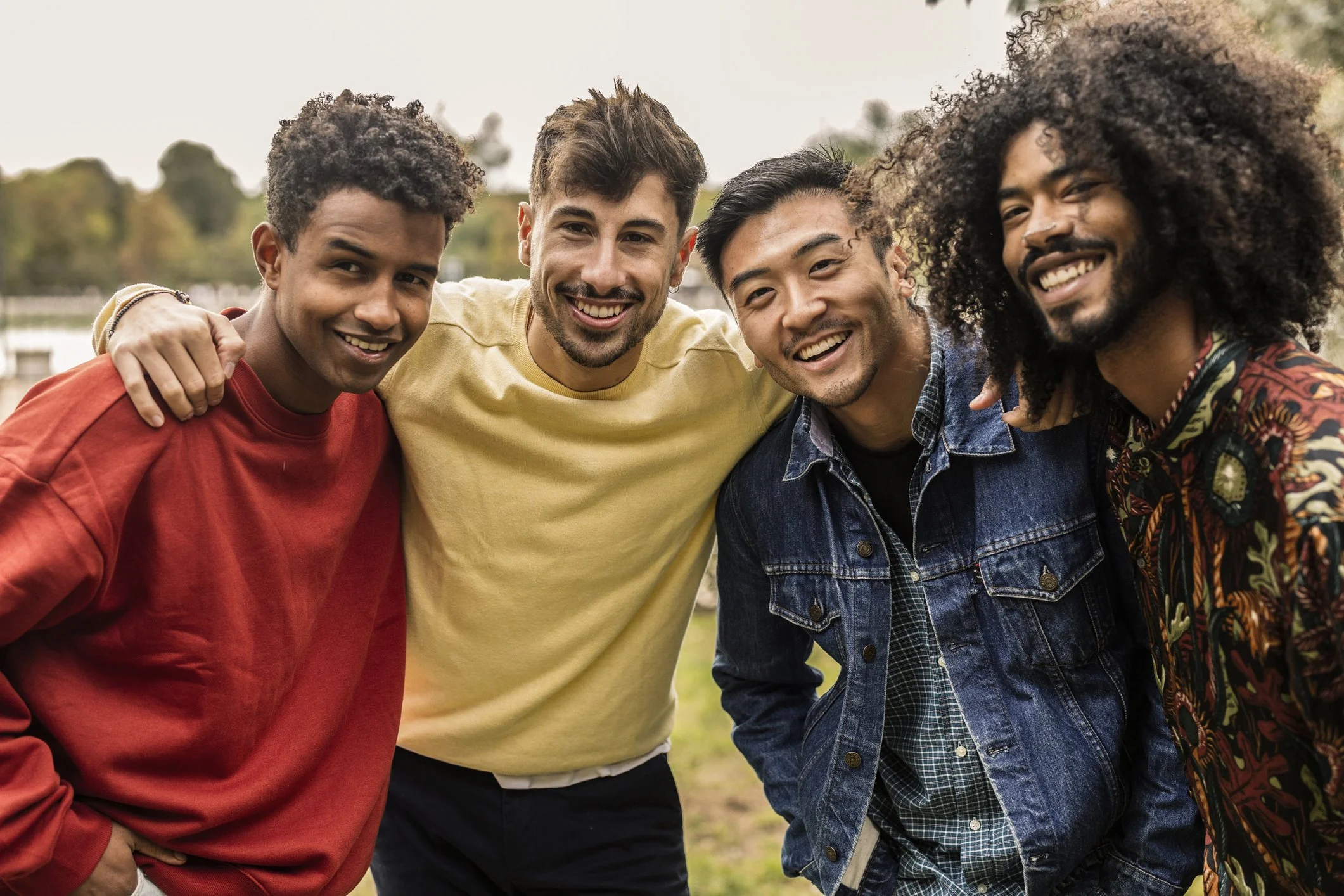Group of five young men with different hair styles smiling and standing close together outdoors.