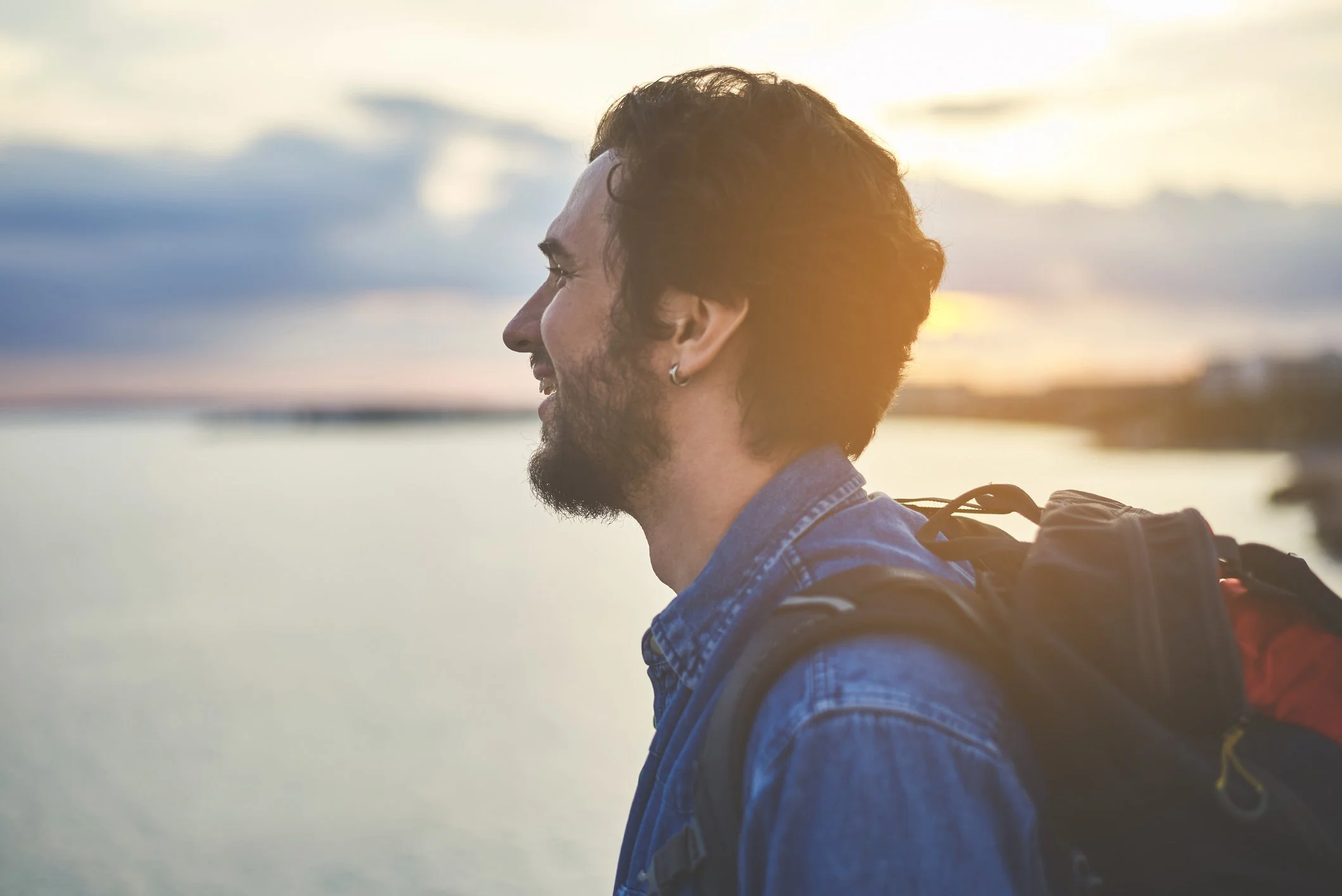 A man with a beard, wearing a denim shirt and earring, smiling naturally, carrying a backpack, standing outdoors near water at sunset.