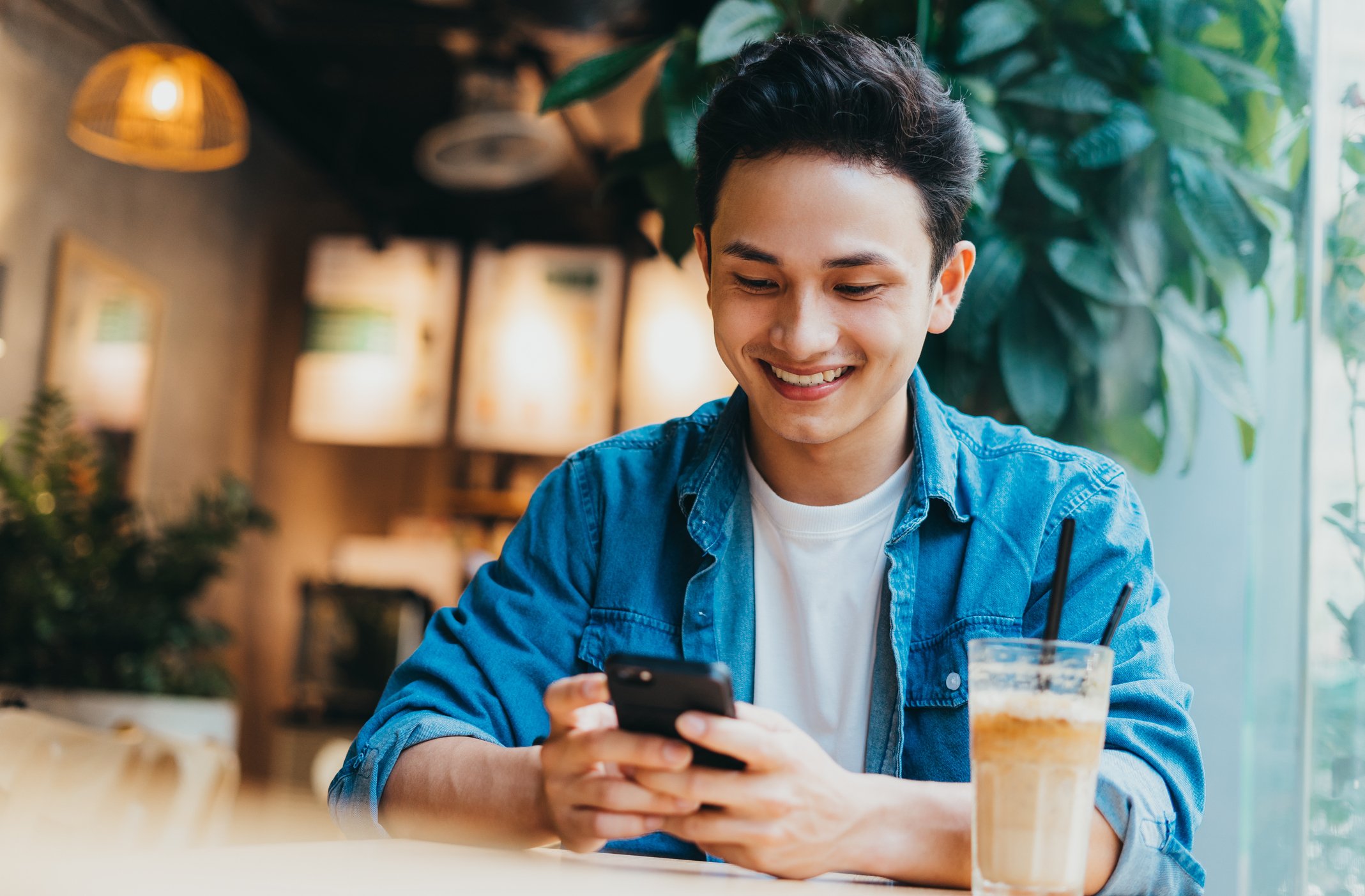 A young man with black hair and a casual blue denim shirt sitting at a table in a cozy, well-lit cafe, smiling while looking at his smartphone. There are two iced coffee drinks on the table in front of him, and a large window with green plants in the background.