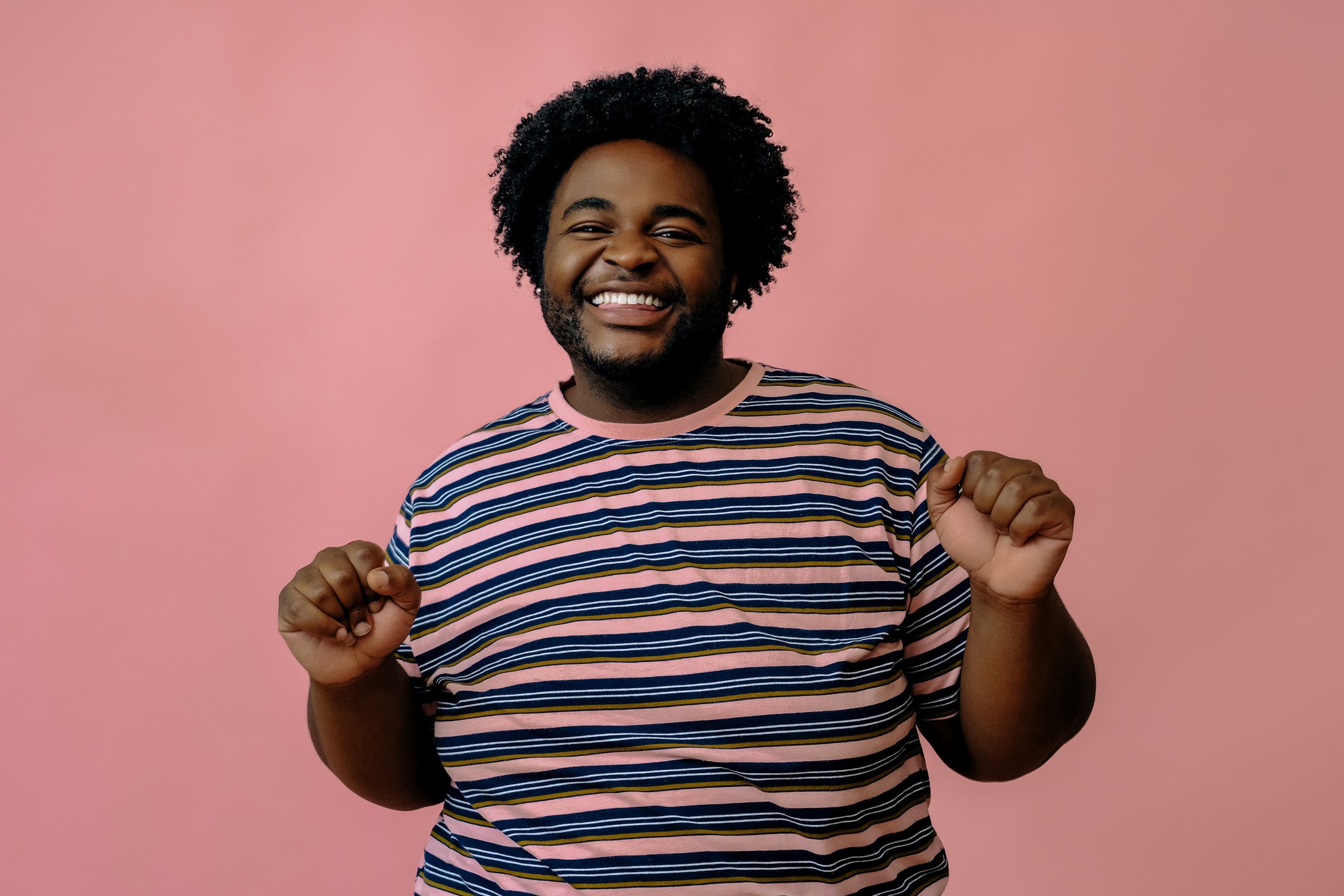 A smiling Black man with curly hair and a beard, wearing a striped t-shirt, standing against a pink background, with his fists raised in a celebratory or triumphant gesture.