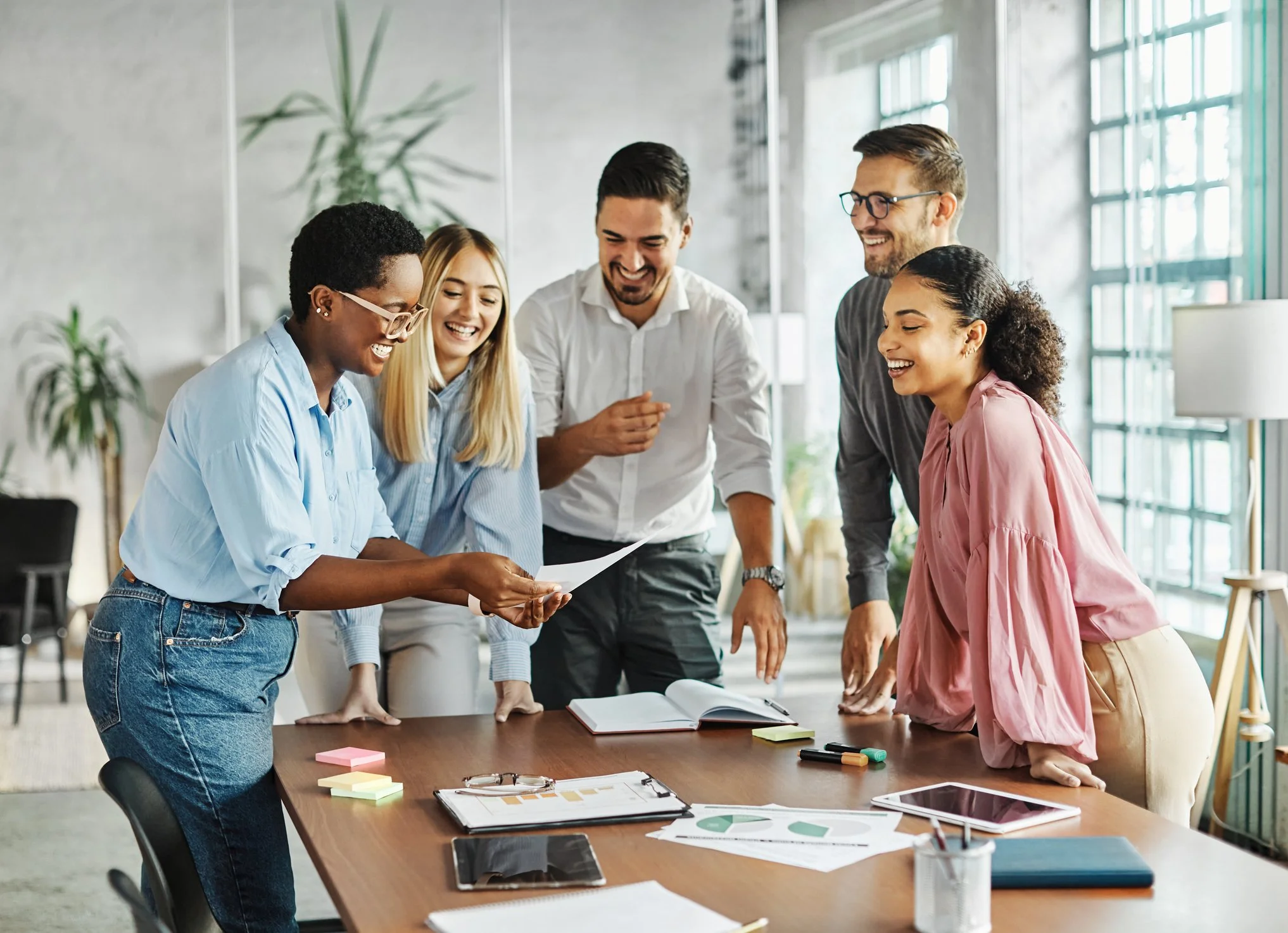 Group of six diverse professionals gathered around a conference table, smiling and discussing documents in a modern office space.