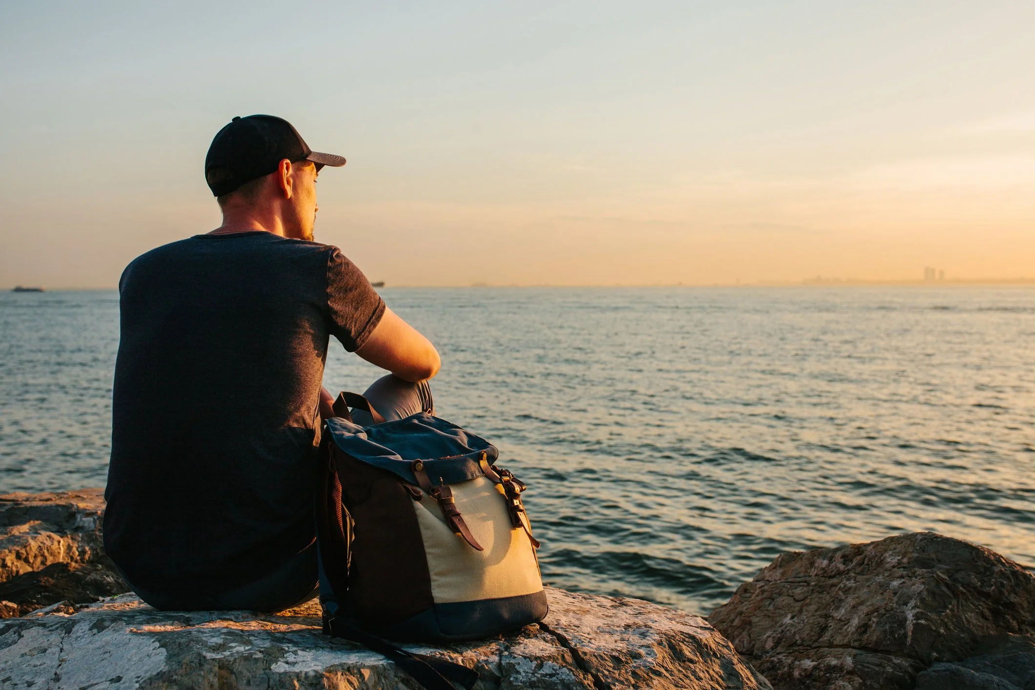 A man sitting on rocks near the water at sunset, wearing a black cap and black t-shirt, with a backpack beside him, gazing at the horizon.