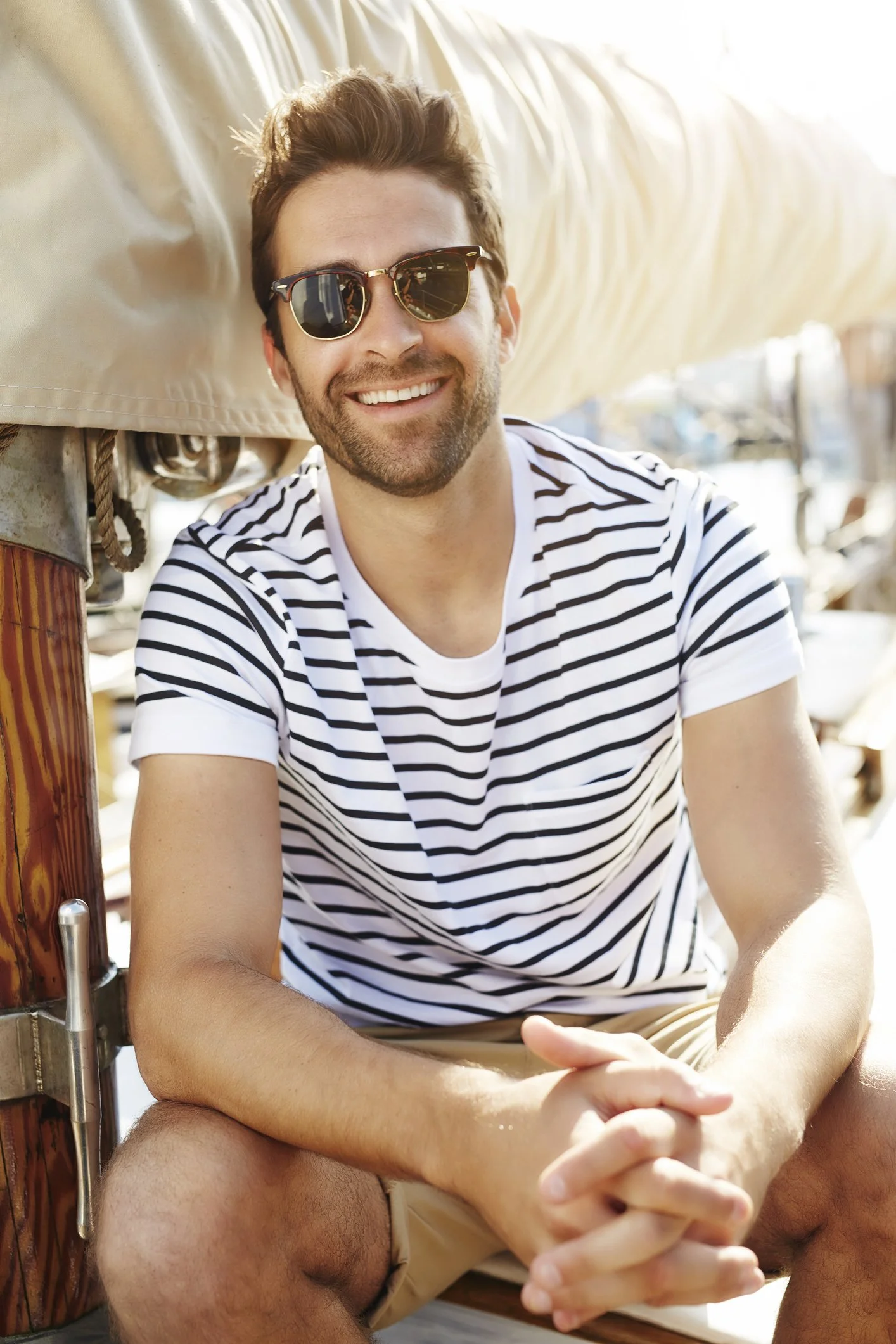A smiling man with brown hair, sunglasses, and a striped shirt sitting outdoors on a sunny day.