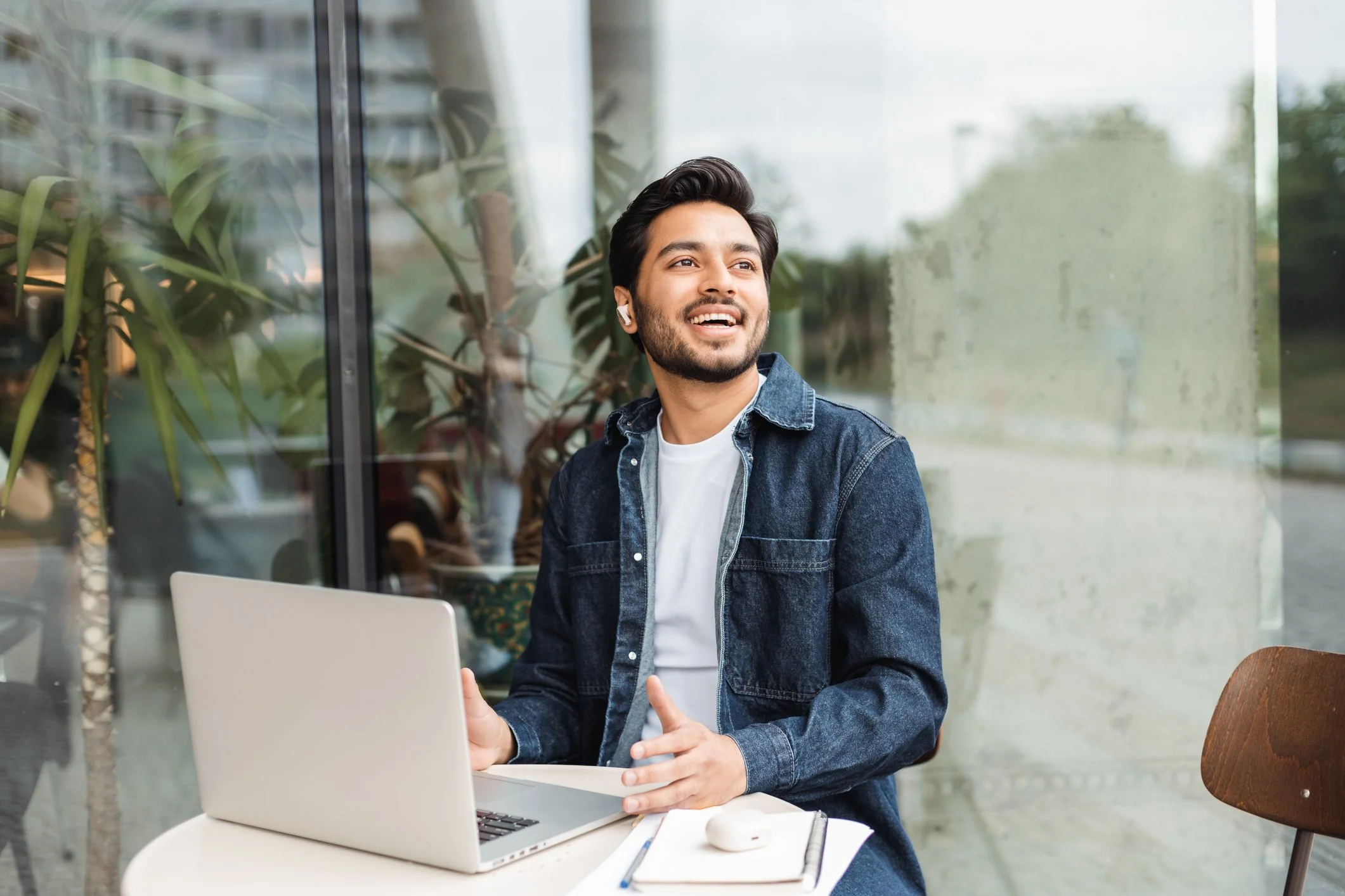 Young man with dark hair, beard, wearing a denim jacket, sitting at a table with a laptop, smiling and wearing wireless earbuds, in a modern indoor space with large windows and green plants visible outside.