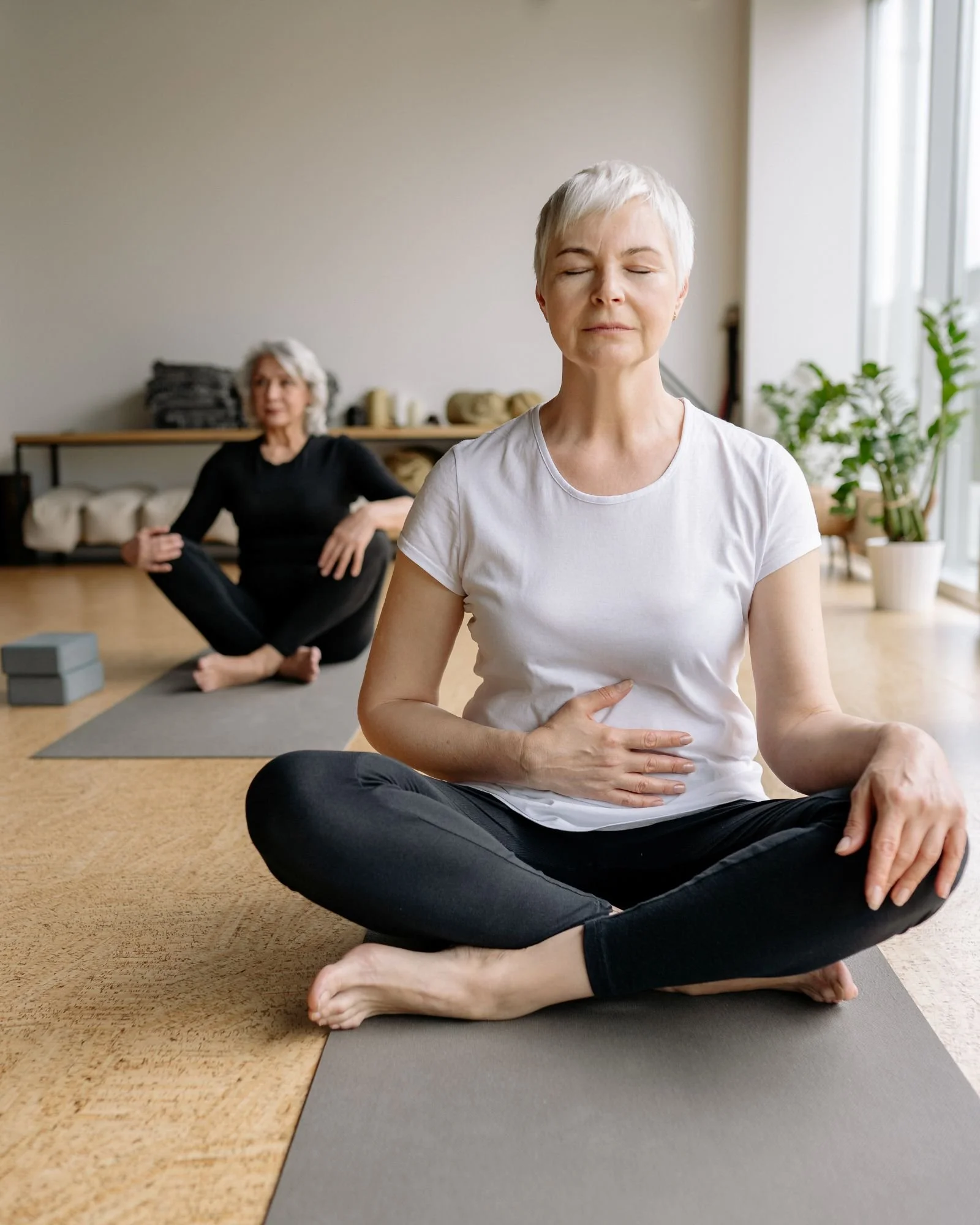 Two mature women seated on yoga mats in a light filled movement studio in Henderson, Nevada, practicing gentle breathwork and mindful movement, focusing on abdominal awareness, nervous system regulation, and long term mobility.