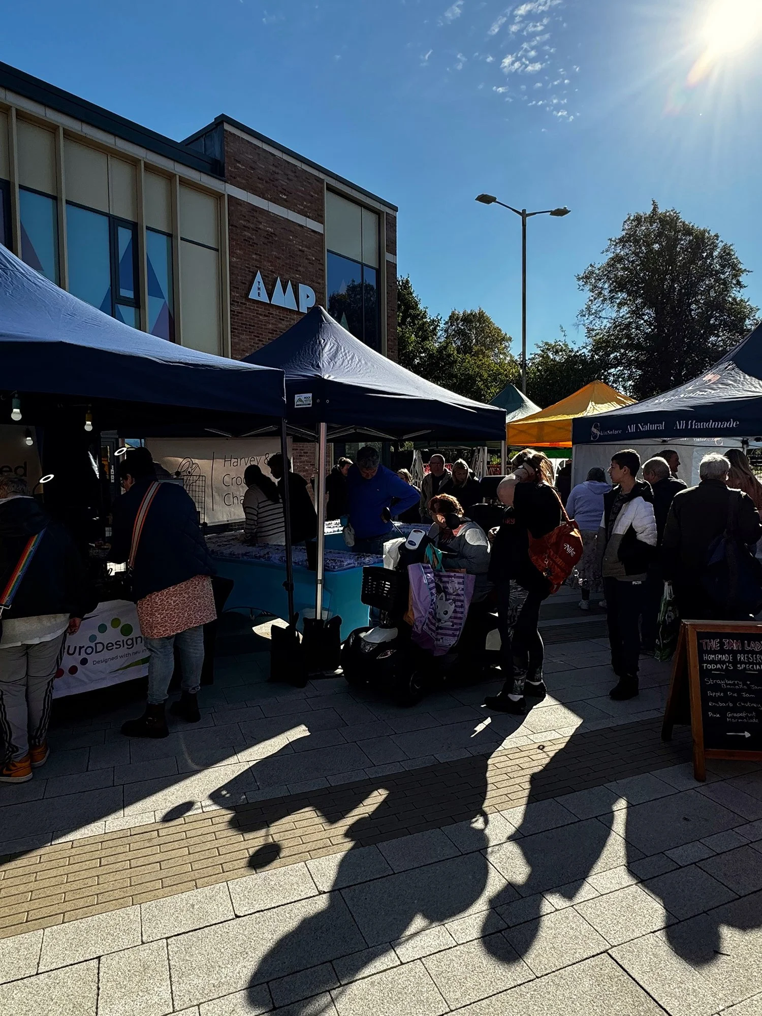 People shopping at outdoor market stalls with tents on a sunny day, outside a building with a sign that reads 'IMAP'.
