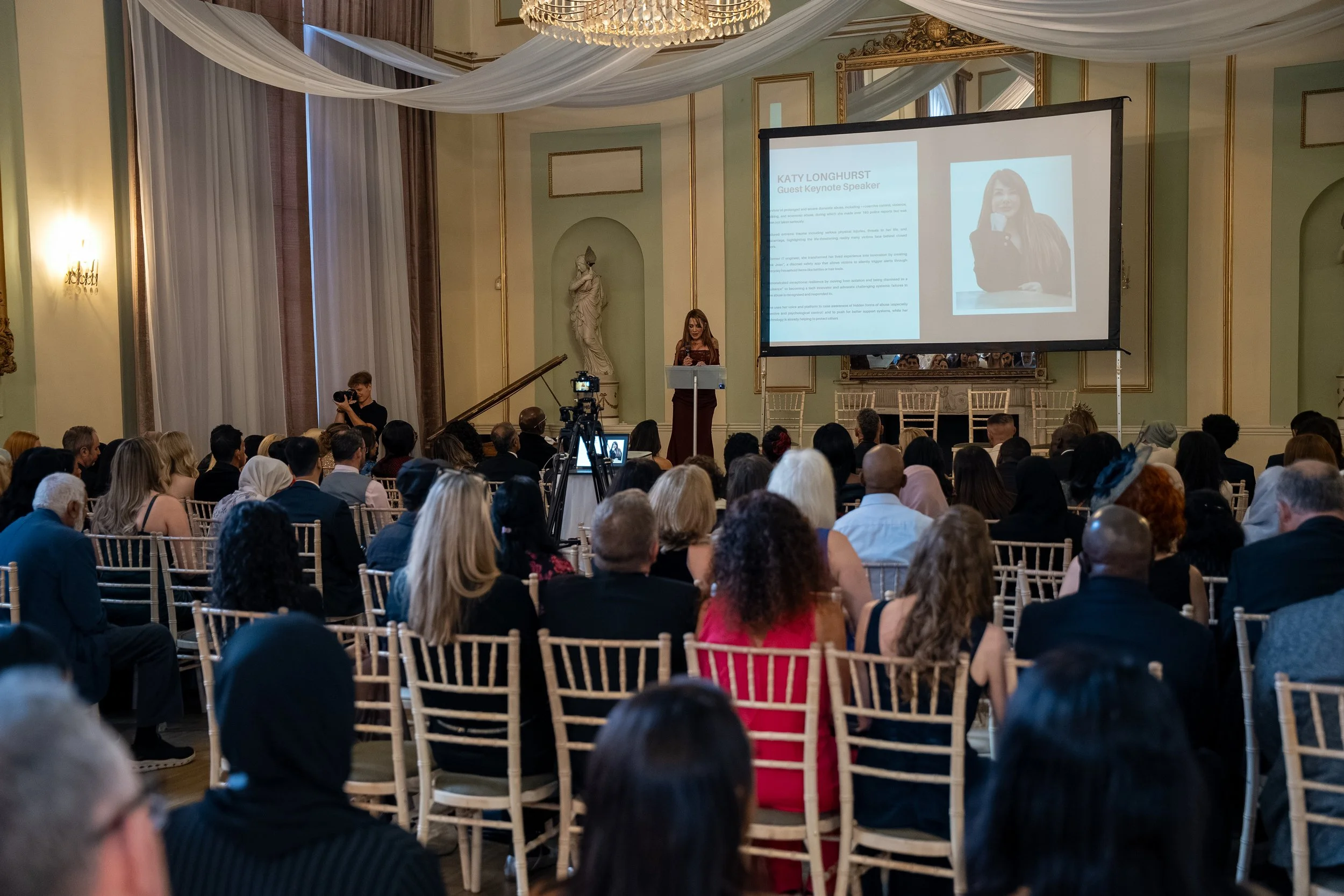 A woman standing at a podium giving a speech in a large, elegant room with chandeliers and white drapery. A presentation slide projects her photo and name, Katy Longhurst, on a large screen. The audience is seated, listening attentively.