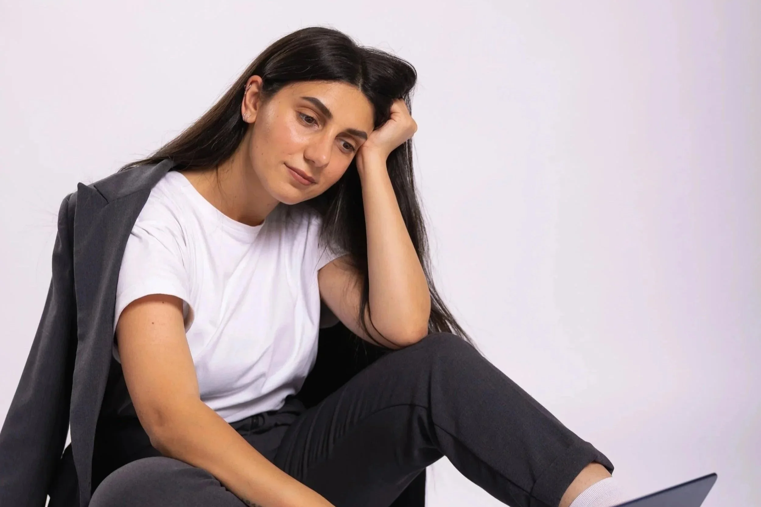 A young woman with long dark hair, wearing a white t-shirt and black pants, sitting and leaning her head on her hand against a plain white background, looking contemplative.