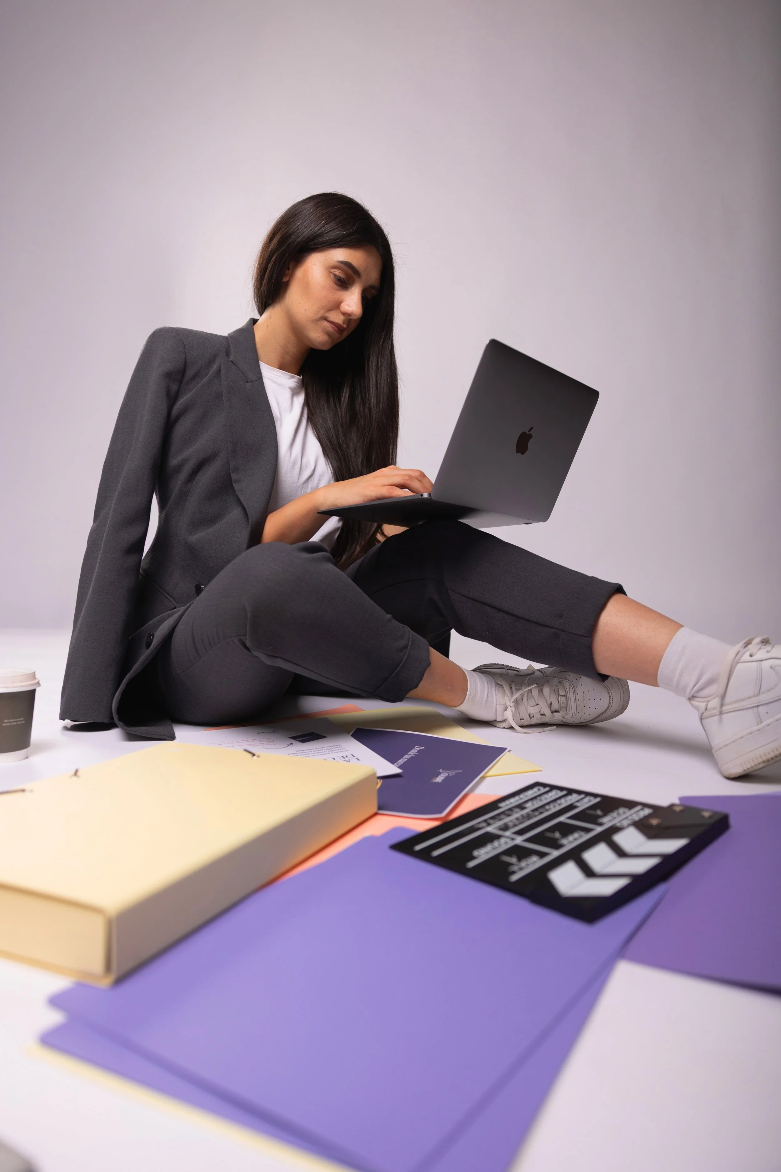A woman with long dark hair, wearing a gray blazer and black pants, is sitting on the floor using a silver MacBook. There are various colored papers, notebooks, and a coffee cup scattered around her on the floor.