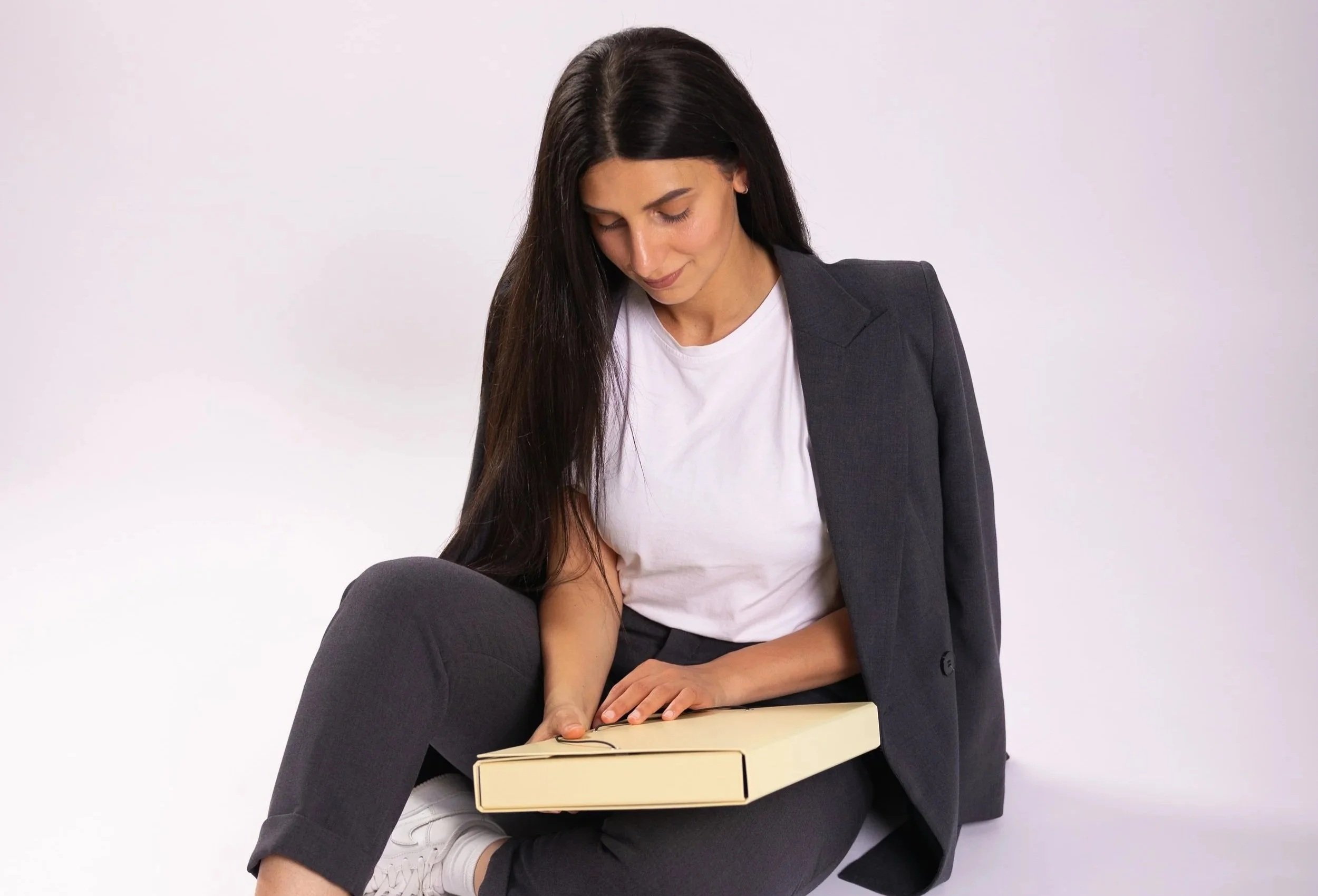 Woman with long dark hair in business attire sitting on the floor; she is looking down at a closed cardboard box she is holding on her lap.