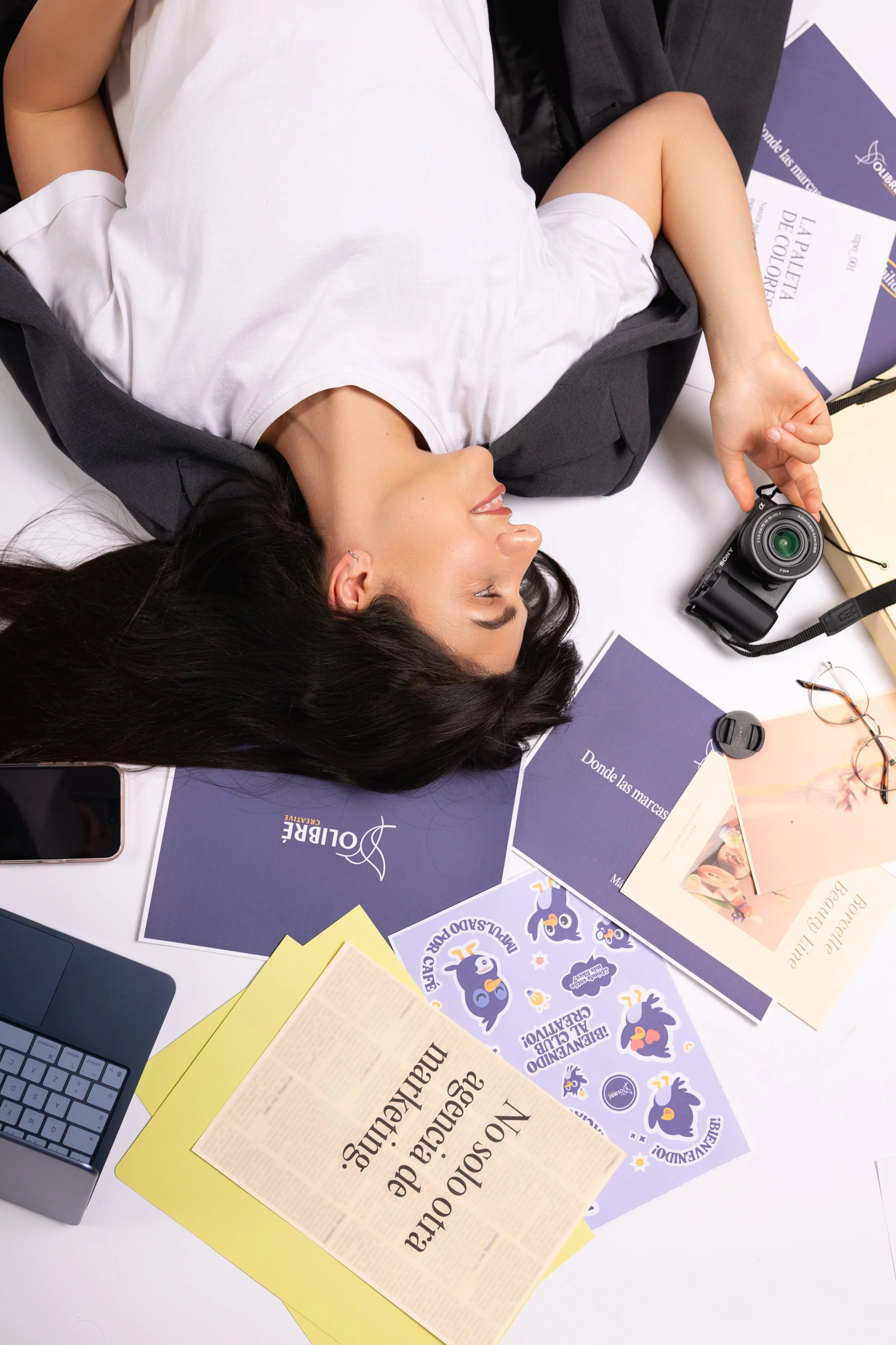 A woman lying on a desk surrounded by brochures, flyers, a camera, a laptop, a smartphone, and glasses, during a work or study session.