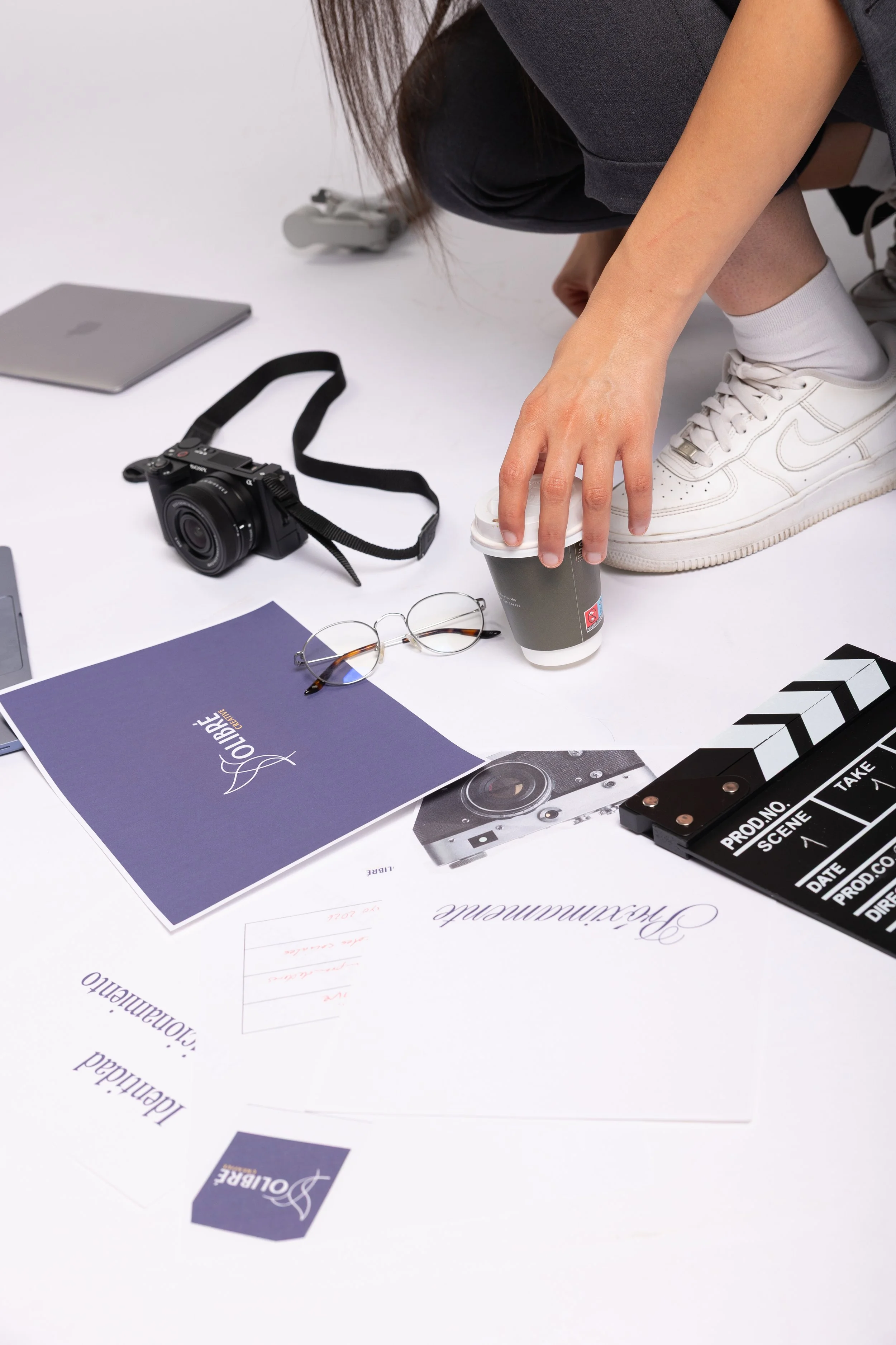 Person crouching on the floor with various objects, including a camera, paper, and a laptop, surrounding her.