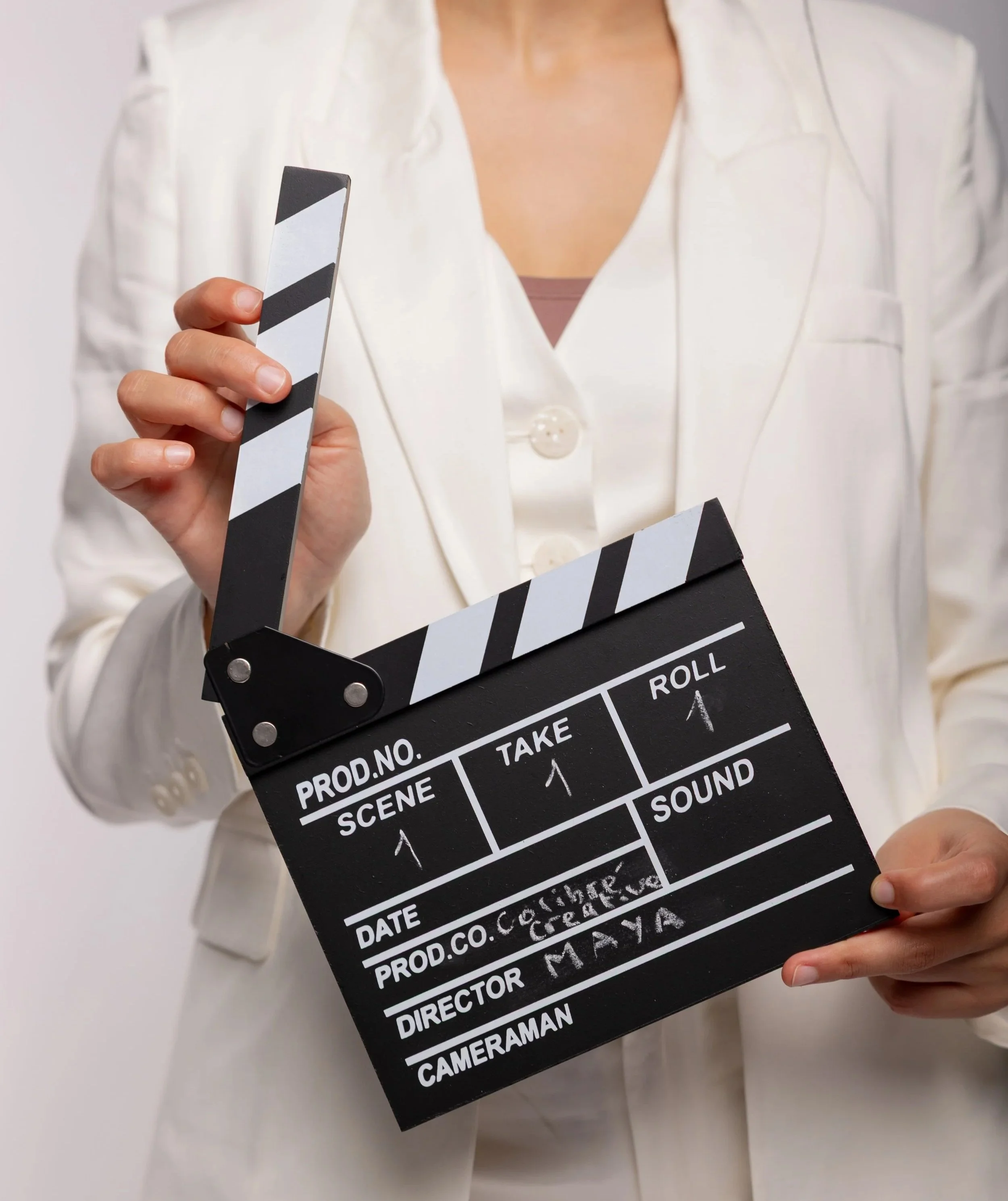 A person dressed in a white blazer holding a black and white clapperboard used in film production, with handwritten notes on it.