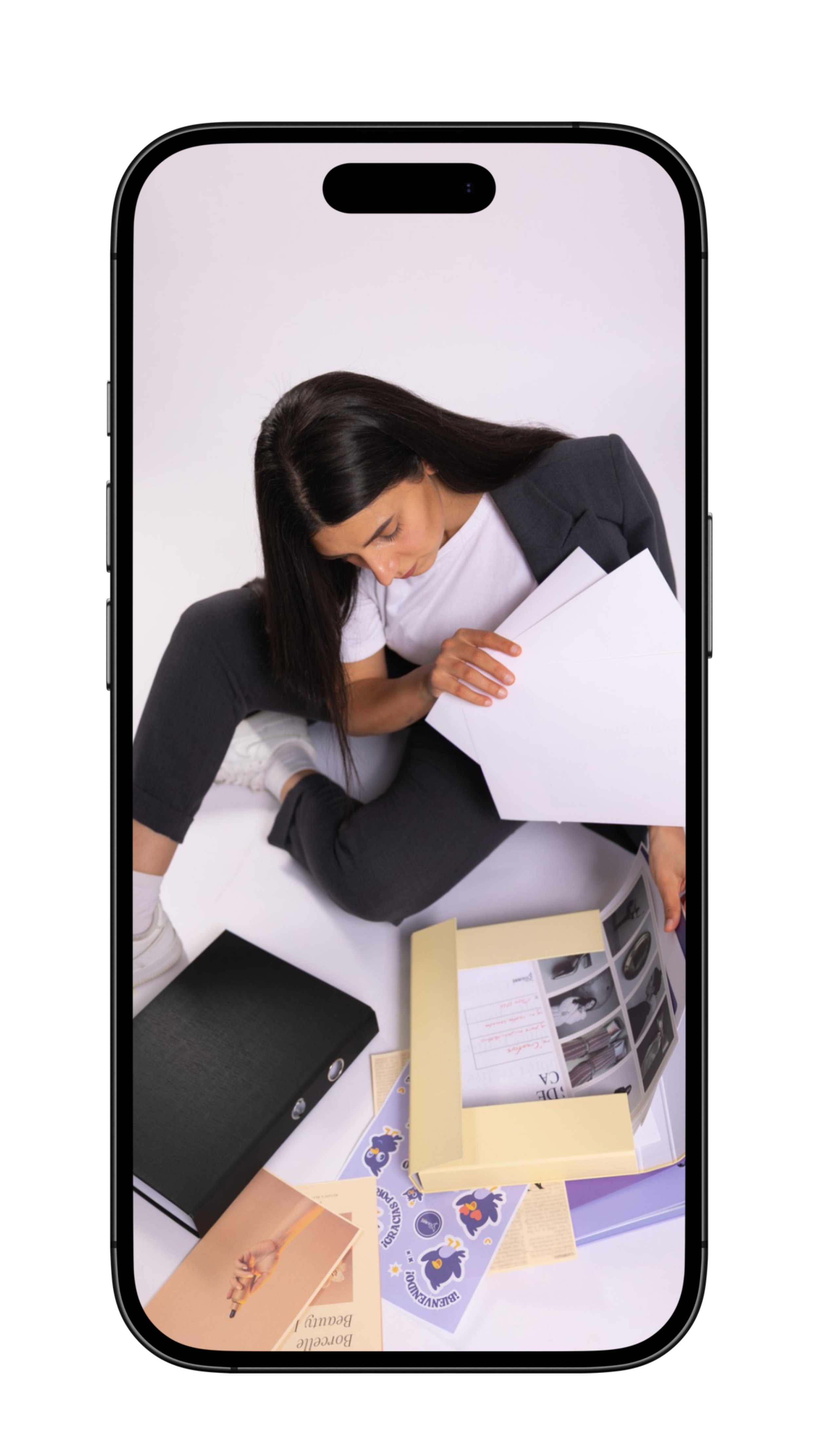 View of a person sitting on a white surface surrounded by various papers, notebooks, and magazines, with only their head visible leaning forward, hair covering their face, wearing casual clothing.
