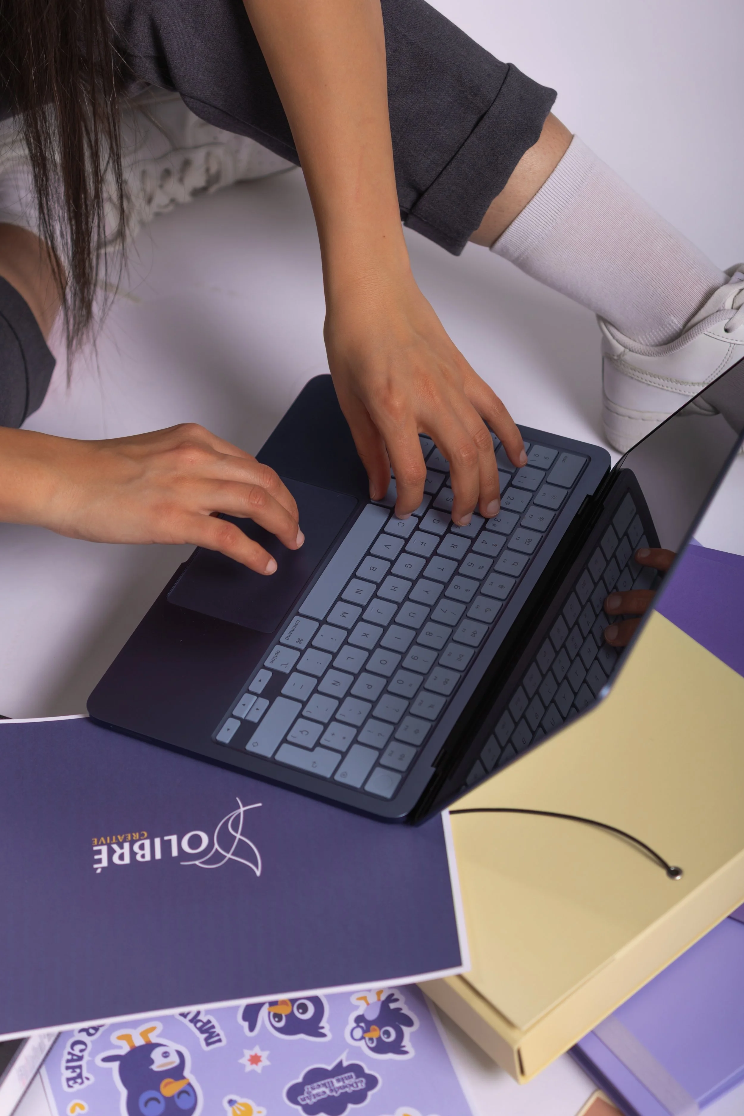 Person sitting on a chair, using a laptop with a blue cover and white keyboard, surrounded by colorful books and papers on a desk.