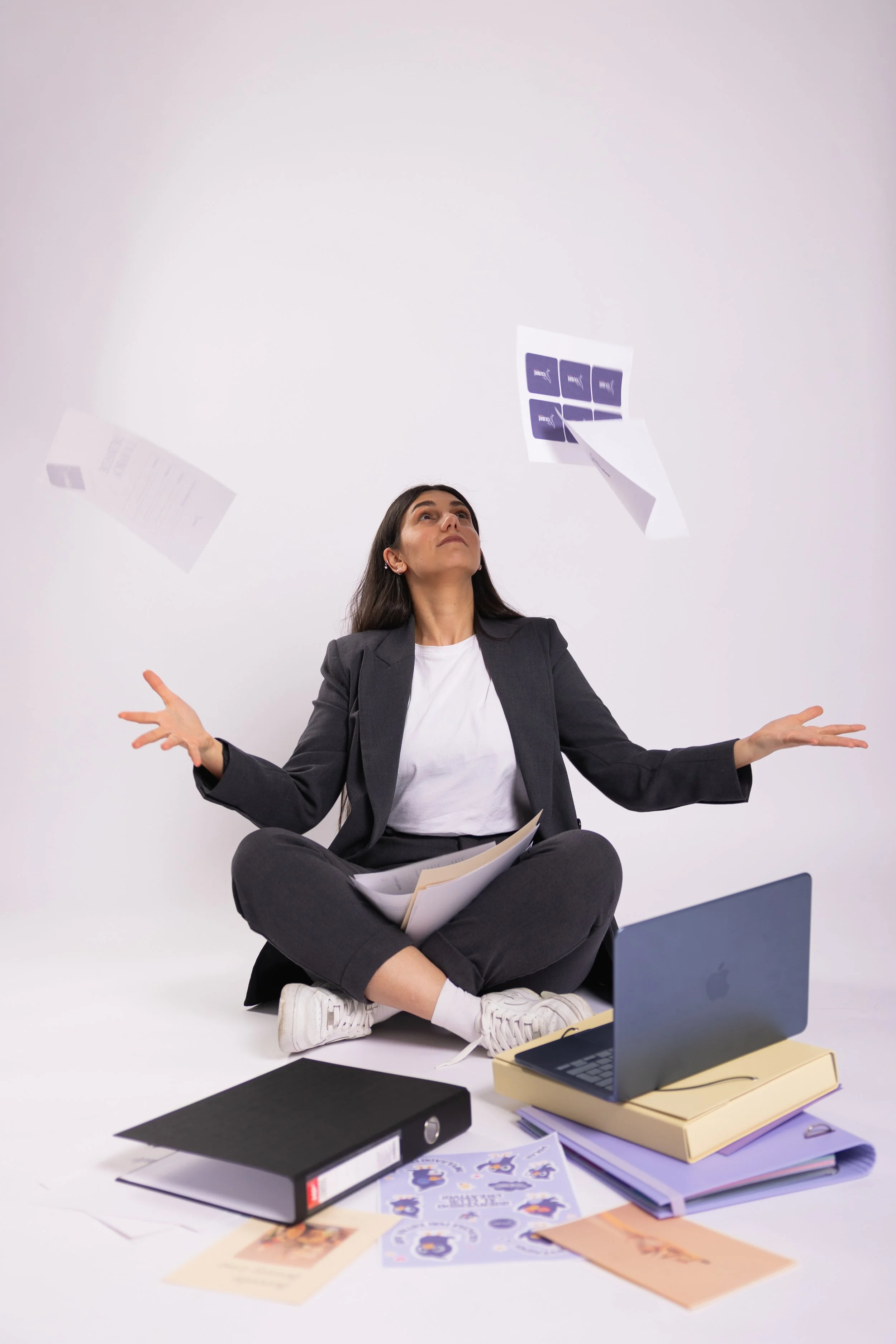 A woman in business attire sitting cross-legged on the floor with papers and folders scattered around her, looking up with a confused or overwhelmed expression. There is a laptop in front of her and papers and stickers flying above.