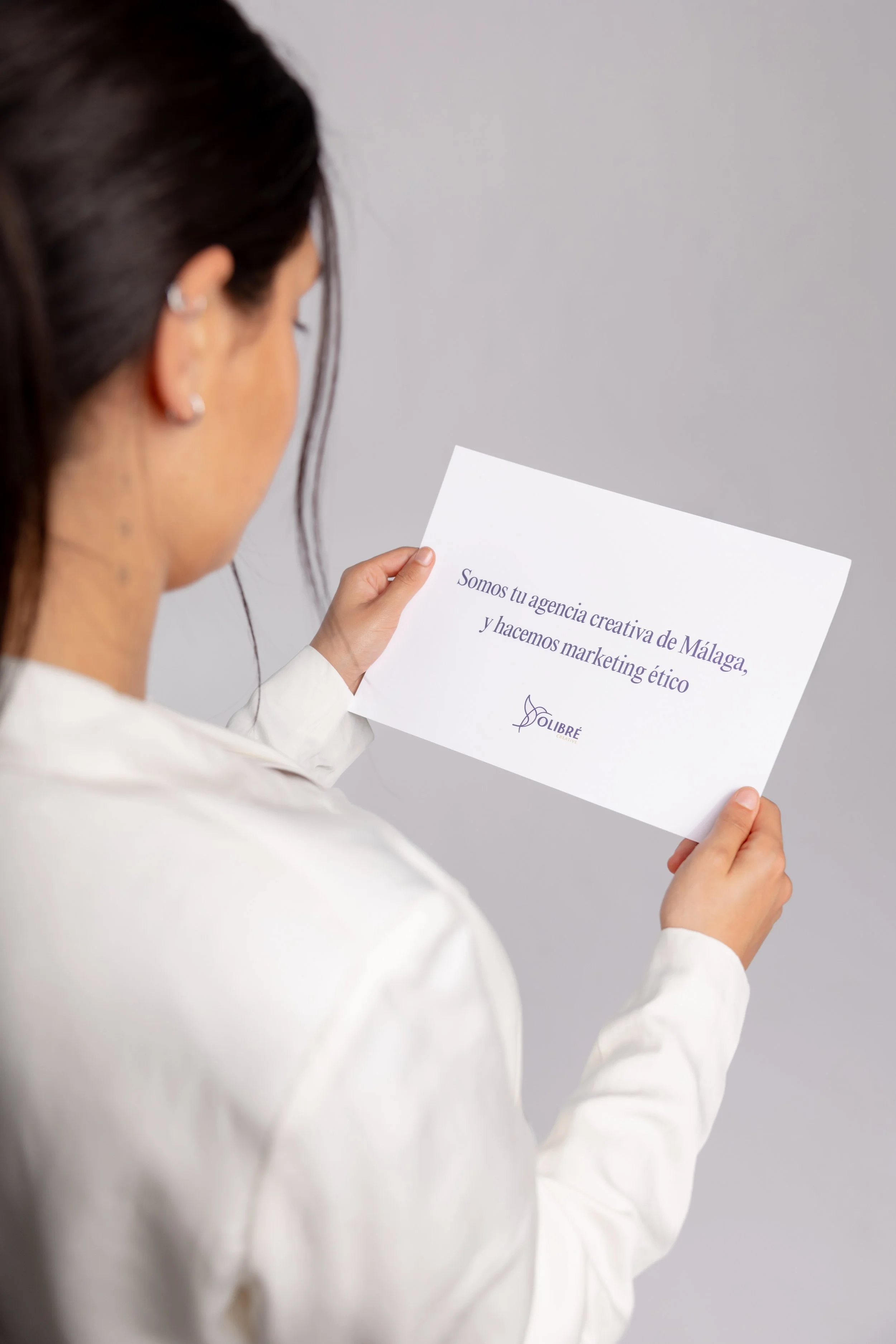 A woman in a white blazer holding a piece of paper with text in Spanish, with her back to the camera and facing a plain gray background.