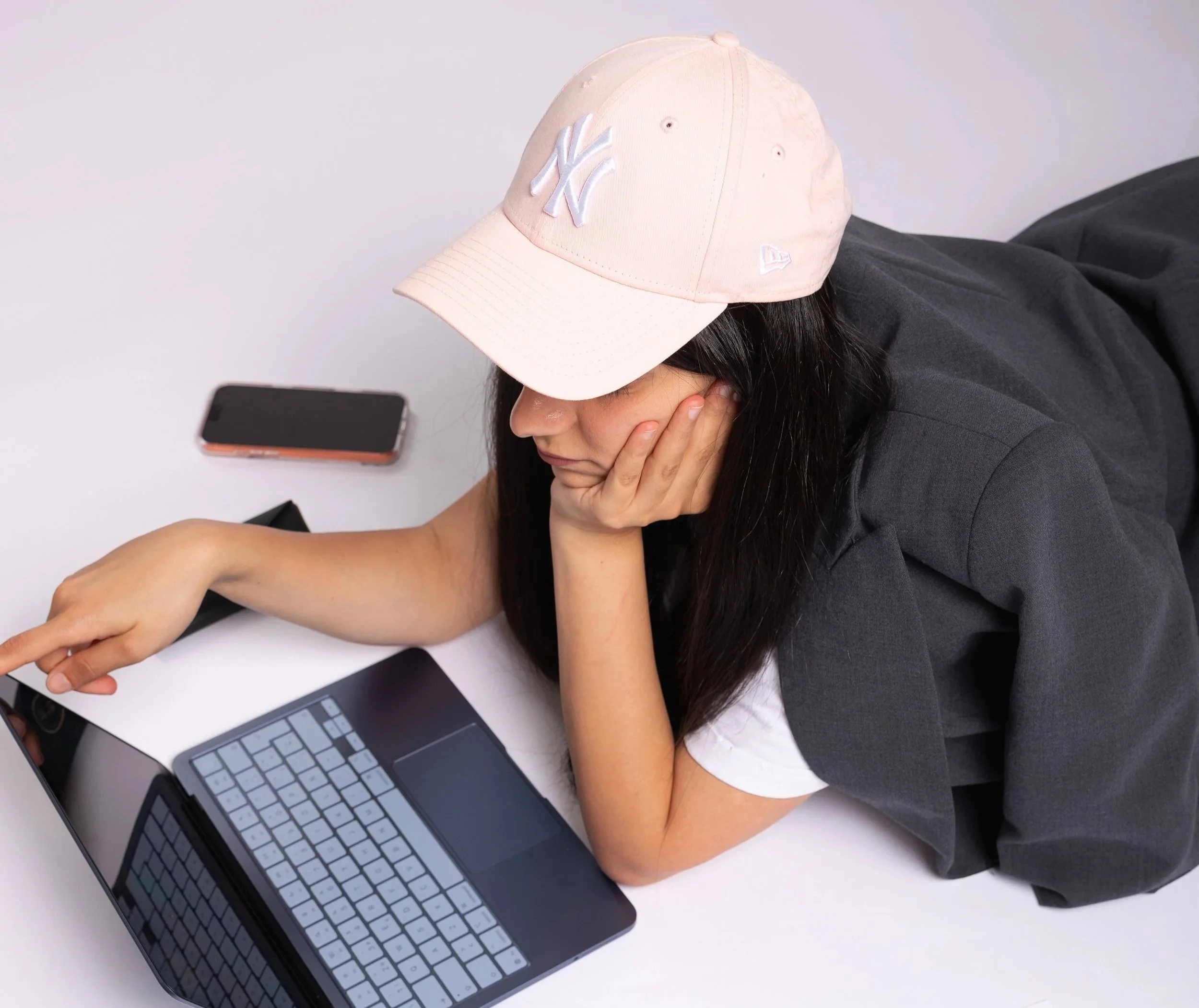 A woman wearing a beige New York Yankees baseball cap and a black shirt with long dark hair, sitting at a white desk with her hand on her face, next to a smartphone and a laptop.