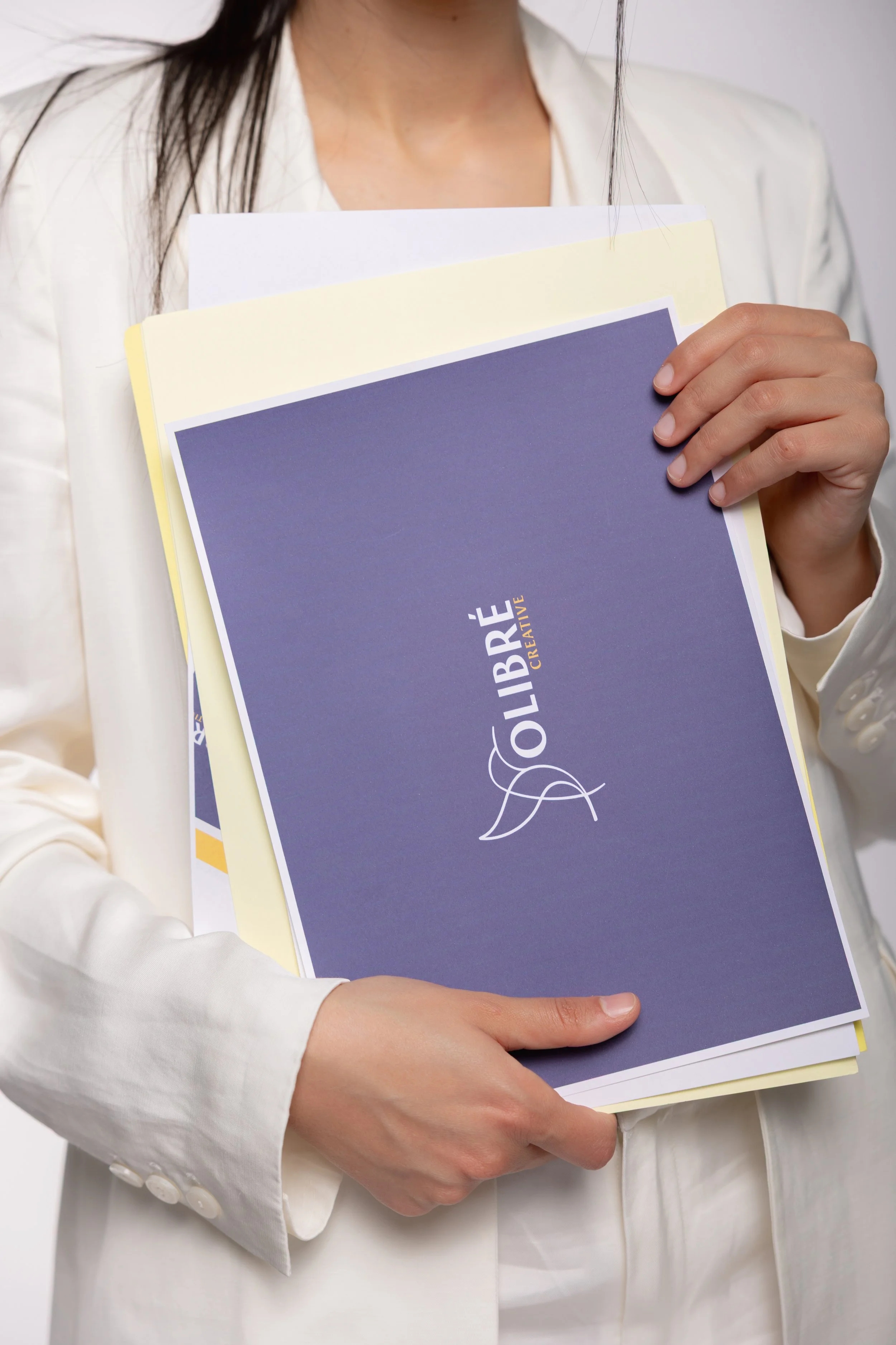 A person wearing a white blazer holding several laminated or printed brochures or folders, with the top one labeled 'LIBRÉ' in large letters and a decorative leaf design.