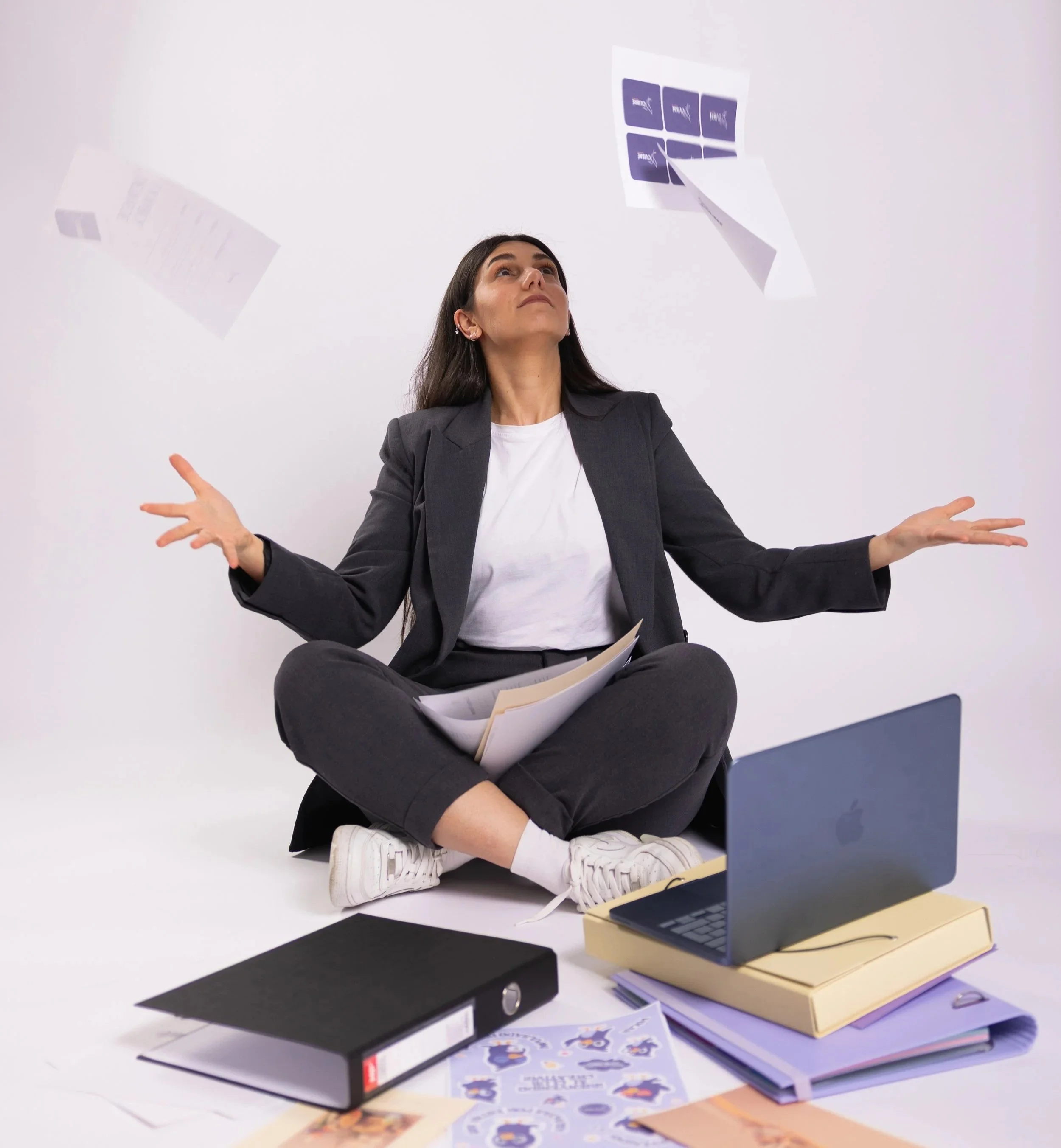 A woman in a gray blazer and white shirt is sitting cross-legged on the floor, juggling papers and folders with a curious expression. There are binders, notebooks, and printed materials spread out in front of her, with some papers flying or falling through the air.