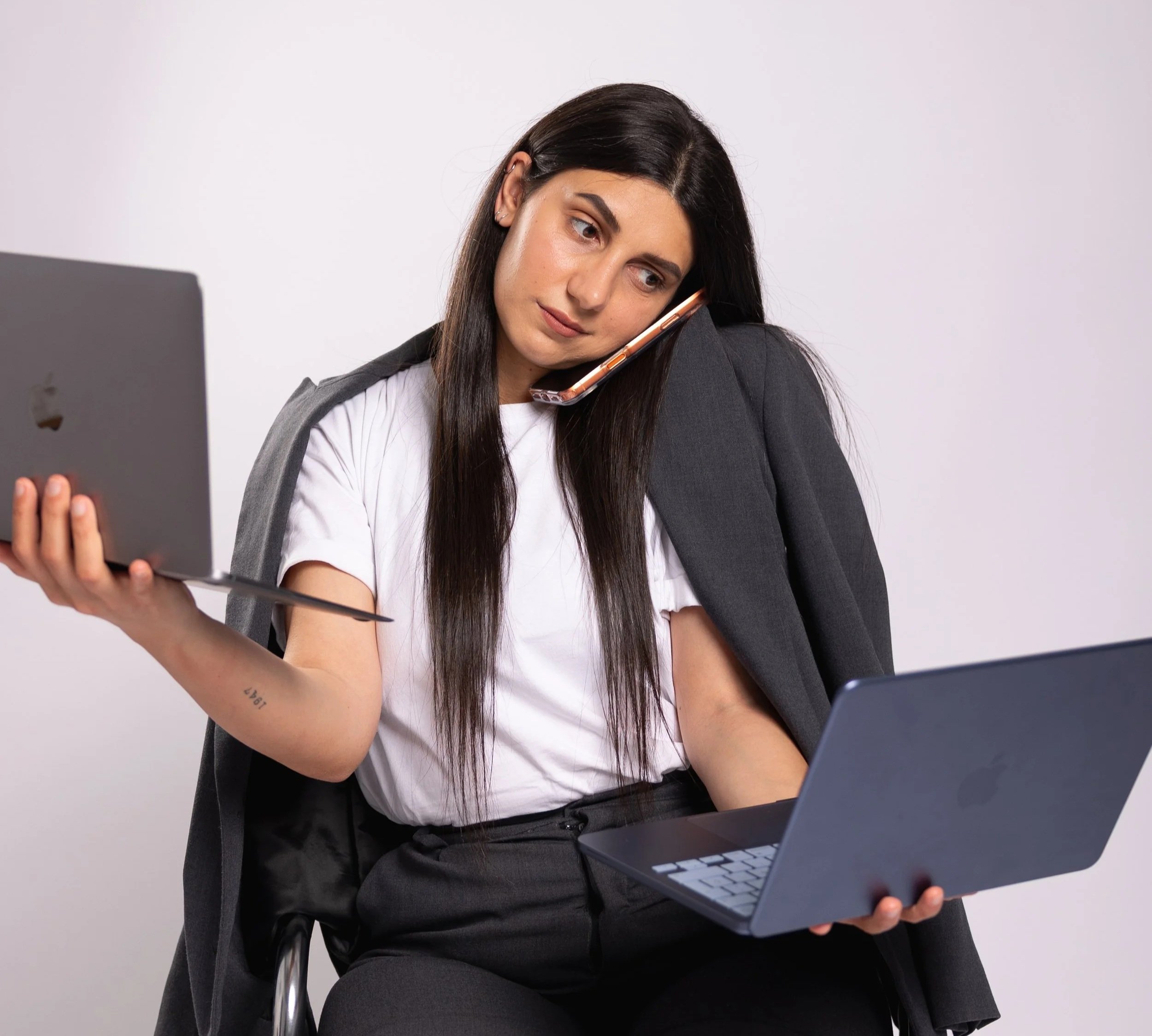 A woman with long dark hair, wearing a white t-shirt and a gray blazer, is sitting and holding a laptop in her left hand and a smartphone pressed against her right shoulder, with a neutral background.