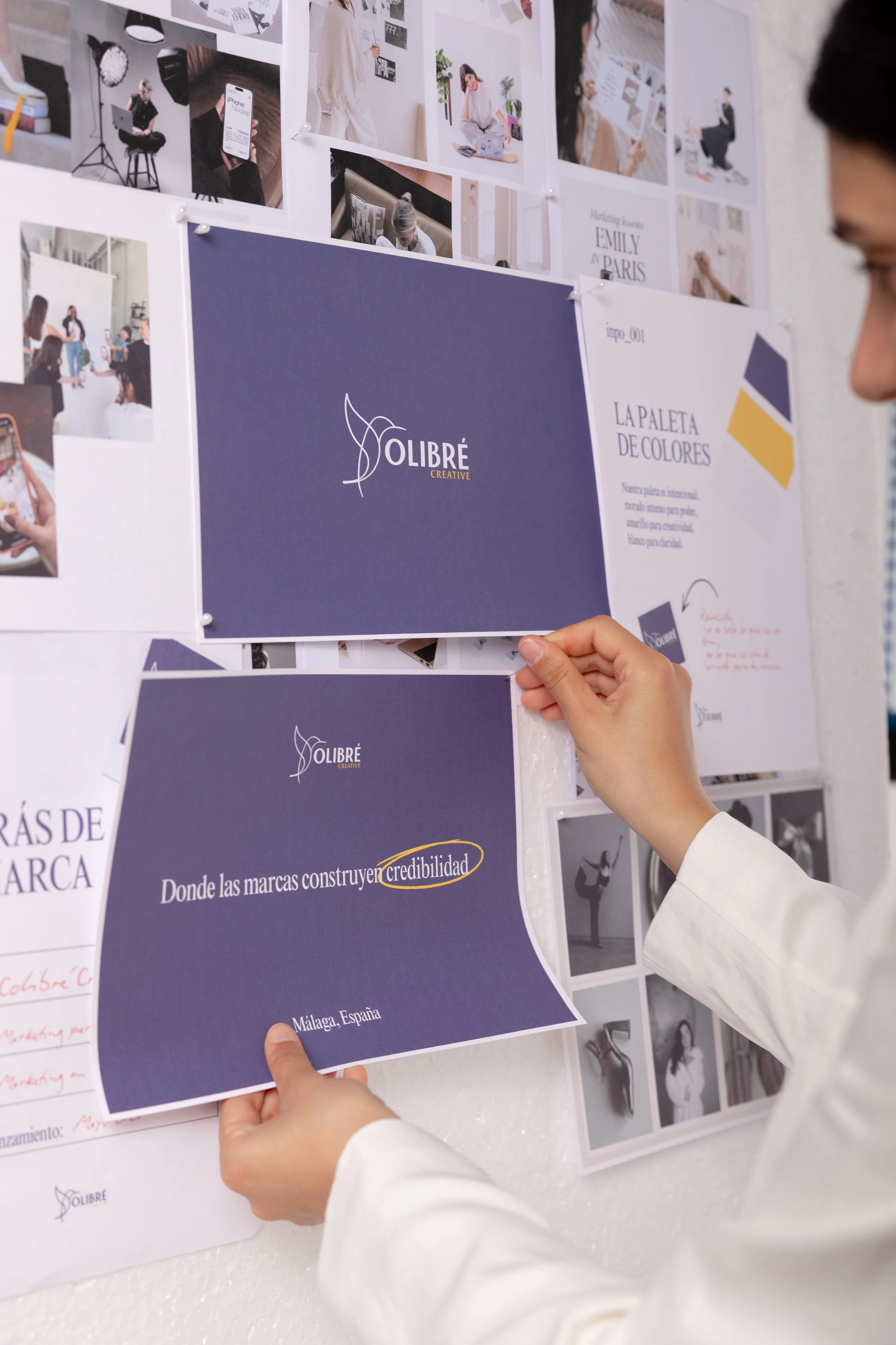 A person in a white shirt holds a blue presentation card that says "LA PALETA DE COLORES" with color swatches, while pinning it to a wall filled with various photographs and design samples.