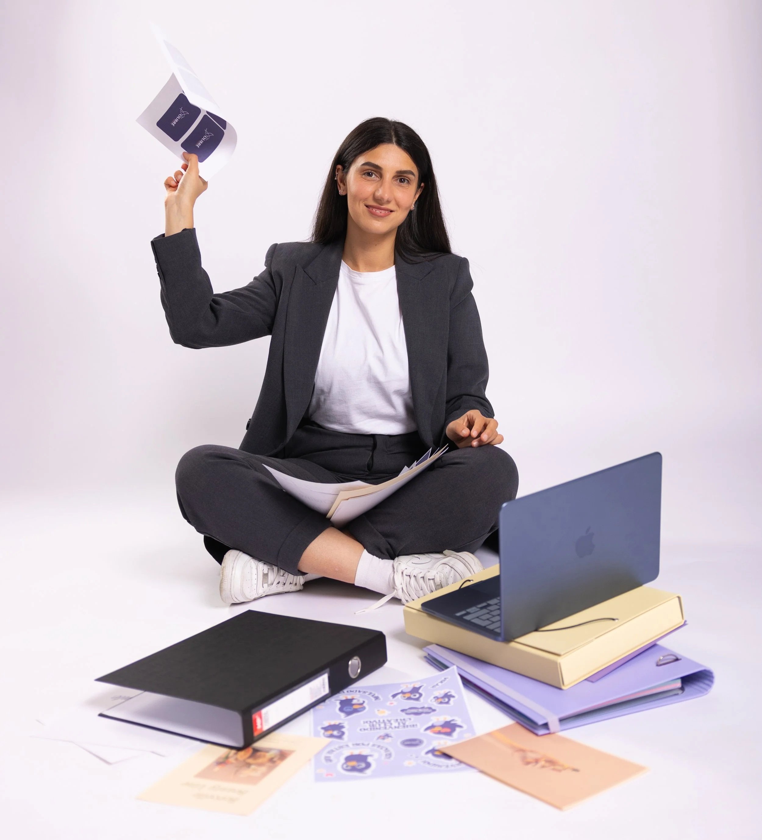 A woman in a grey suit and white t-shirt sitting cross-legged on the floor, smiling, with paperwork, folders, and a laptop in front of her, against a plain white background.