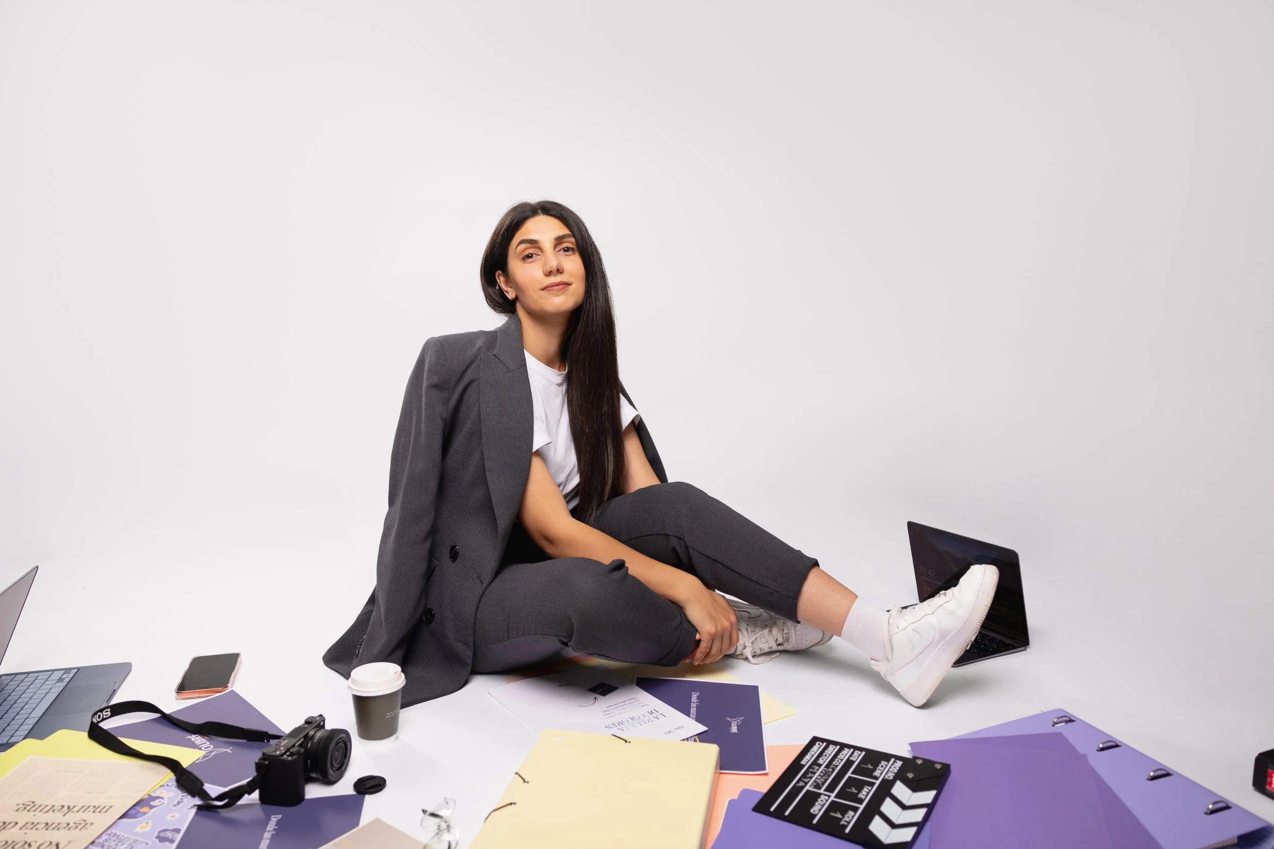 A woman with long dark hair wearing a gray business suit sitting on the floor surrounded by scattered office supplies, including notebooks, papers, a camera, coffee cup, and laptop, on a white background.