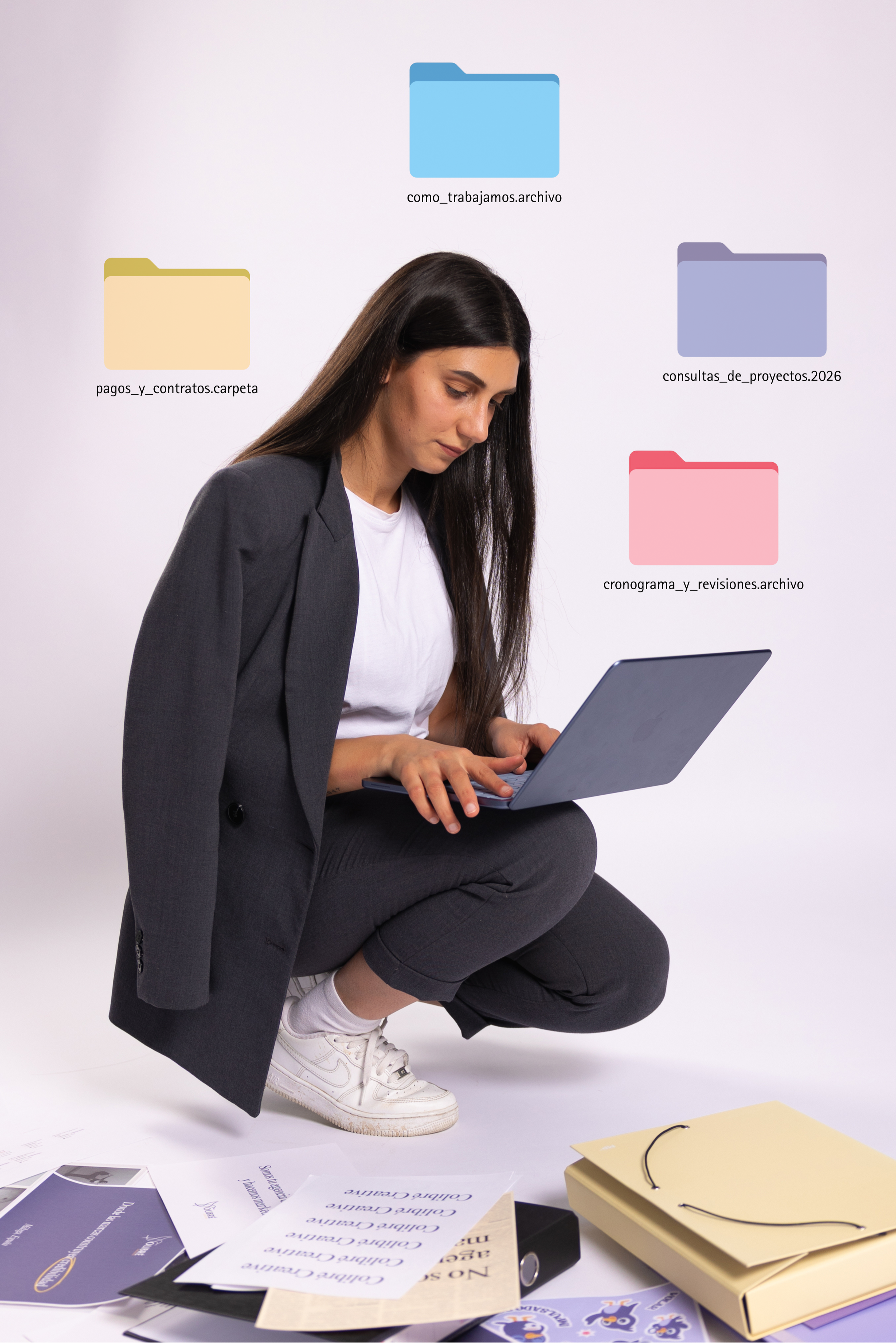 A woman with long dark hair, dressed in a gray blazer and white shirt, is squatting and looking at a laptop. There are scattered papers and brochures on the floor, including a yellow folder and a black binder. The papers contain text and images, some arranged neatly, others overlapping.