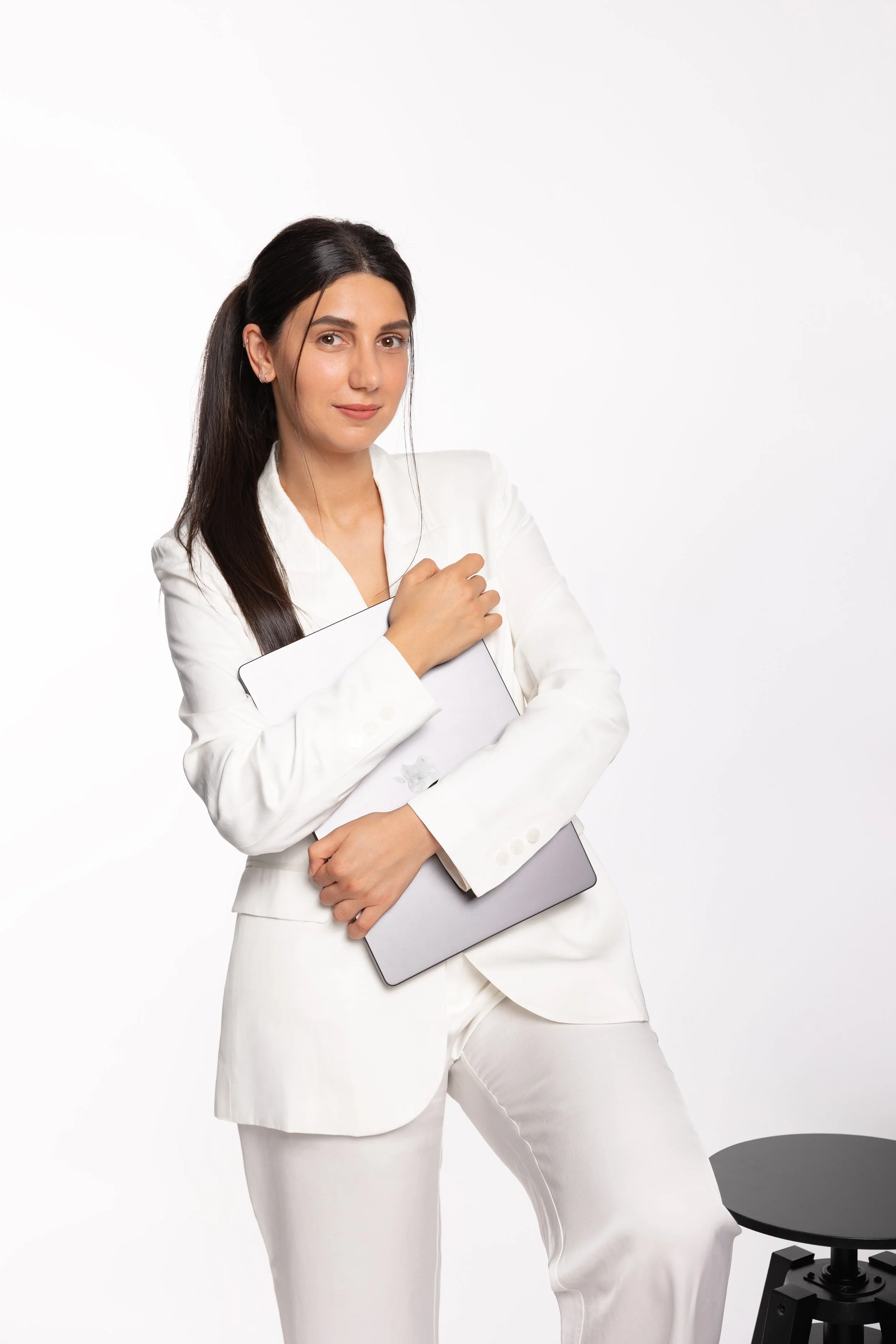 A woman with long dark hair wearing a white suit, holding a gray tablet, standing against a white background.
