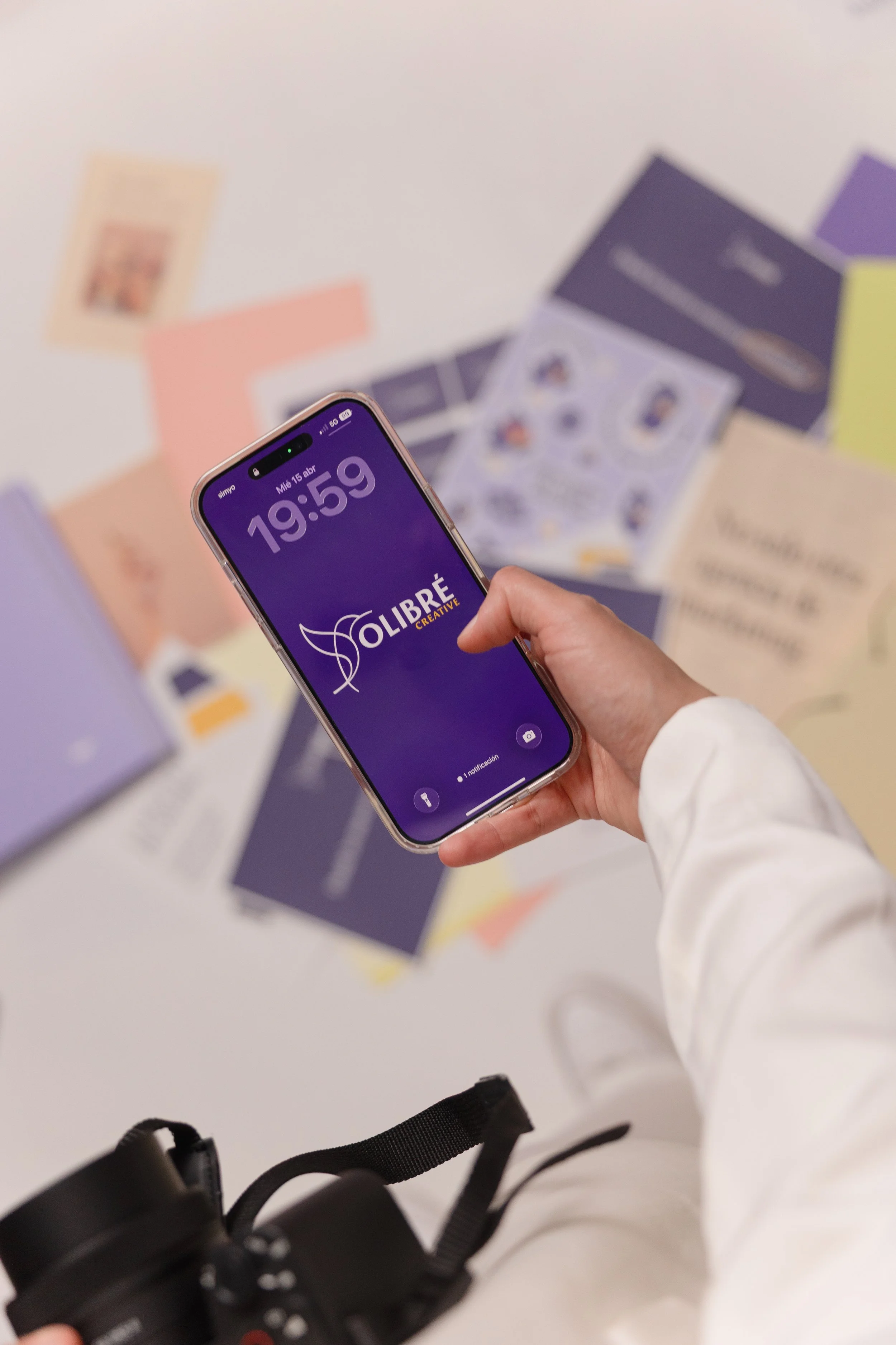 A person holding a smartphone with the time 19:59 on the screen, displaying the logo of OLIBRÉ CREATIVE. The background shows various colorful bookmarks and a camera strap.