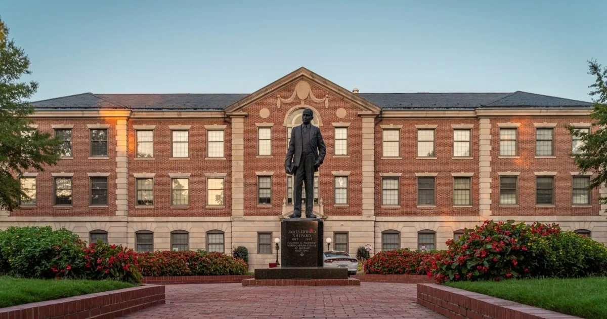 Statue of a man in front of a brick building with large windows and decorative architectural features, surrounded by landscaped gardens and paved walkways at North Carolina Central University.